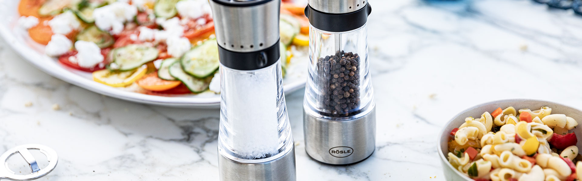RÖSLE Gewürzmühlen und Gerichte Gewürzmühlen aus Edelstahl mit transparentem Behälter, neben einem bunten Salat und einer...