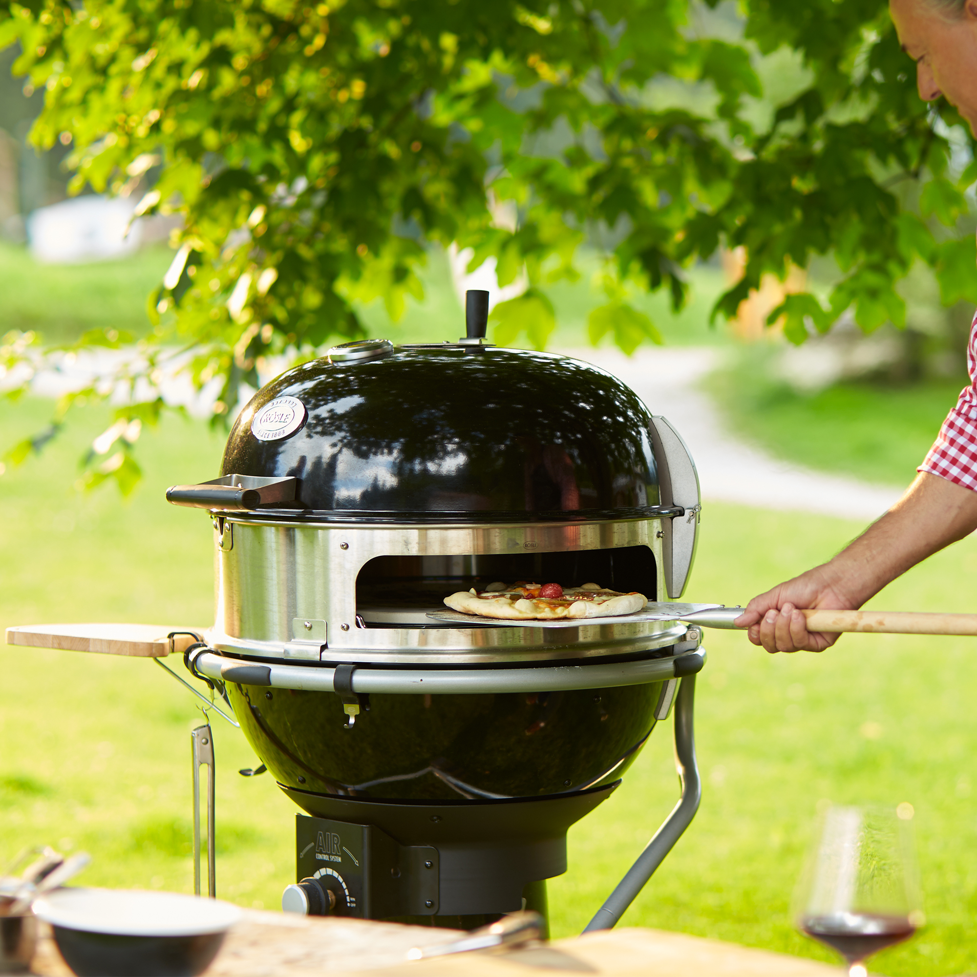RÖSLE Gourmetring No. 1 Grill im Freien Gourmetring No. 1, runder Grill mit schwarzem Gehäuse, im Freien verwendet, Pizza wird herausgenommen.