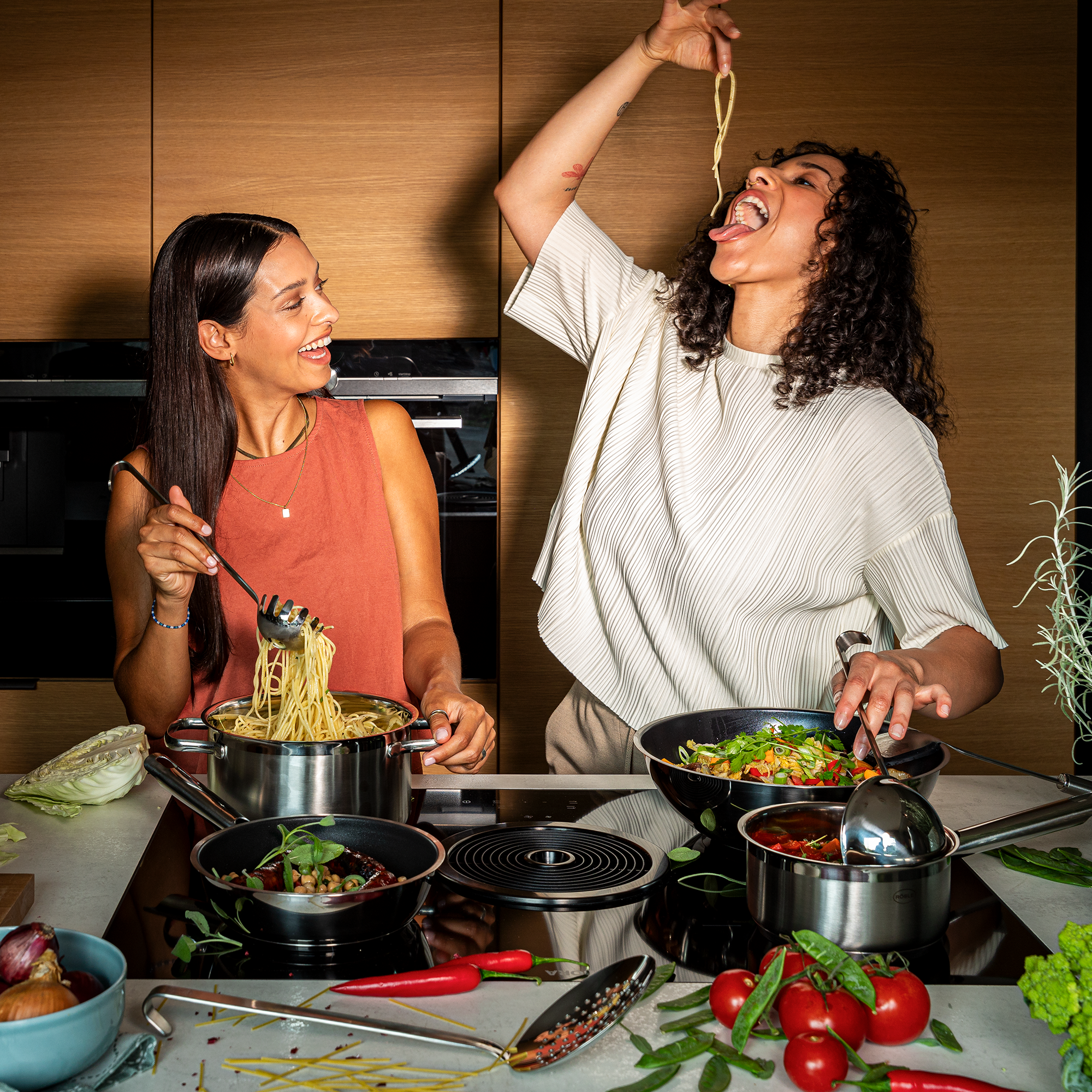 Zwei Frauen in einer modernen Küche, eine hält einen Topf mit Spaghetti, die andere isst mit Freude einen langen Spaghetti-Faden.