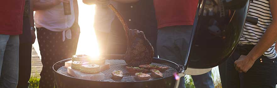Holzkohlegrill von RÖSLE bei Grillfest Holzkohlegrill mit Fleisch und Gemüse auf dem Rost, umgeben von Personen bei einem Grillfest.