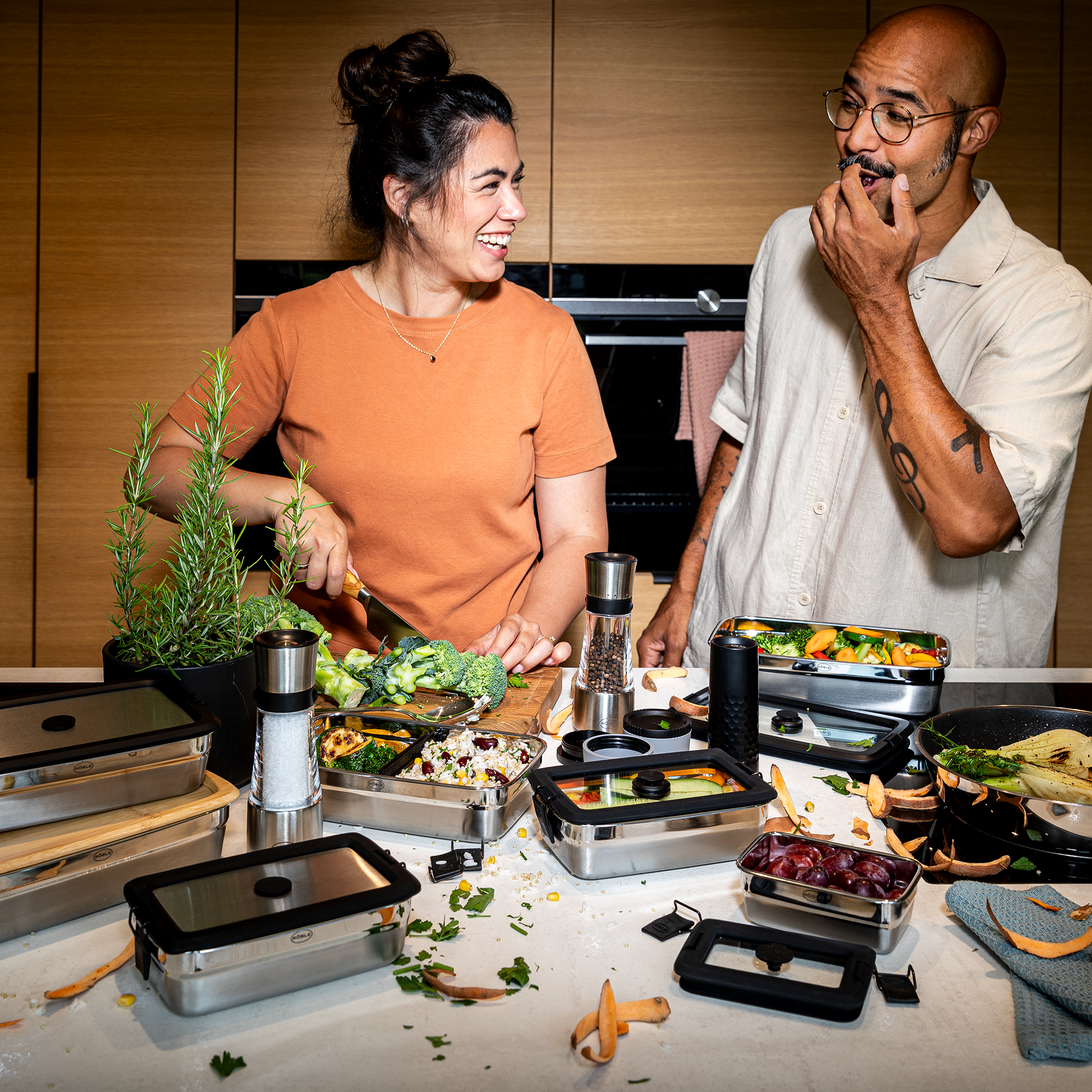 Zwei Personen in der Küche mit RÖSLE Flexx2go Behältern Zwei Personen in einer modernen Küche, eine Frau schneidet Gemüse, der Mann lächelt und hält...