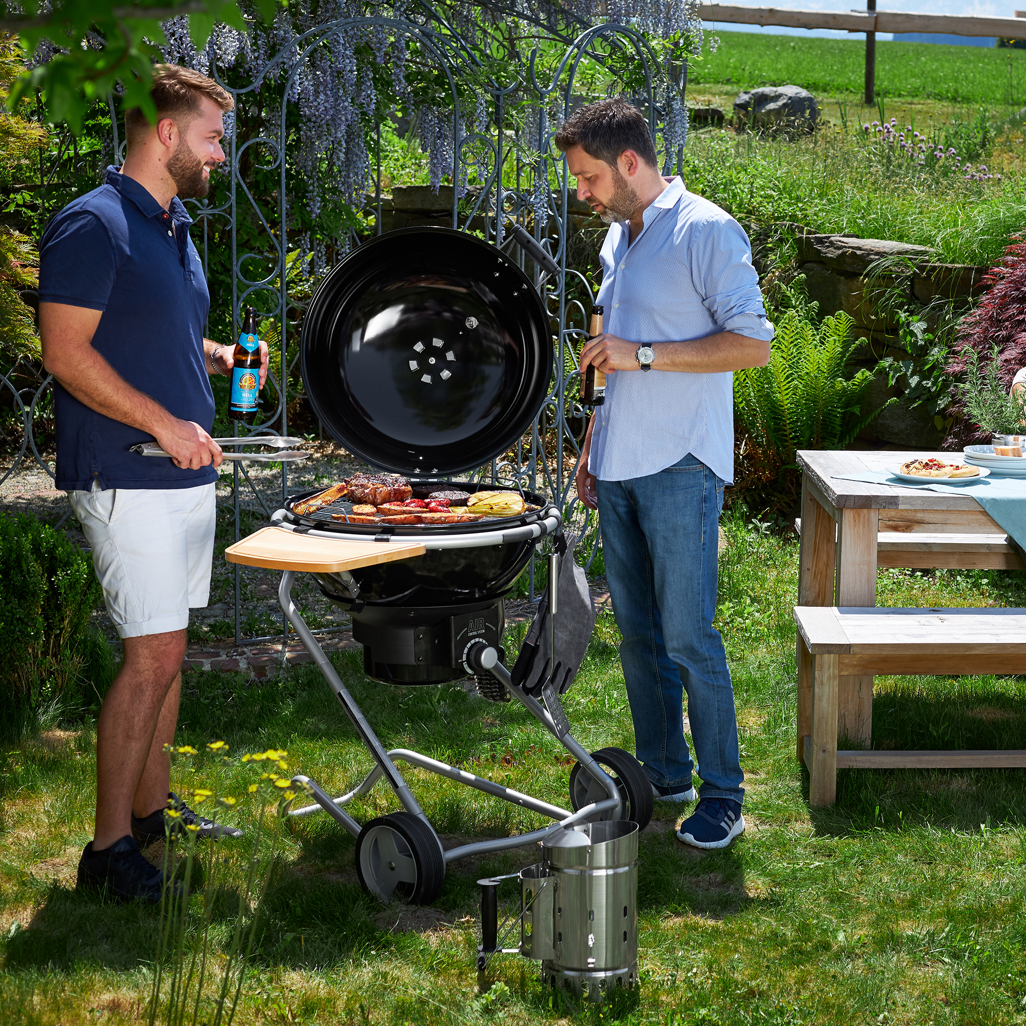 RÖSLE charcoal kettle barbecue in use Two men barbecue with a charcoal kettle barbecue outdoors, surrounded by greenery.