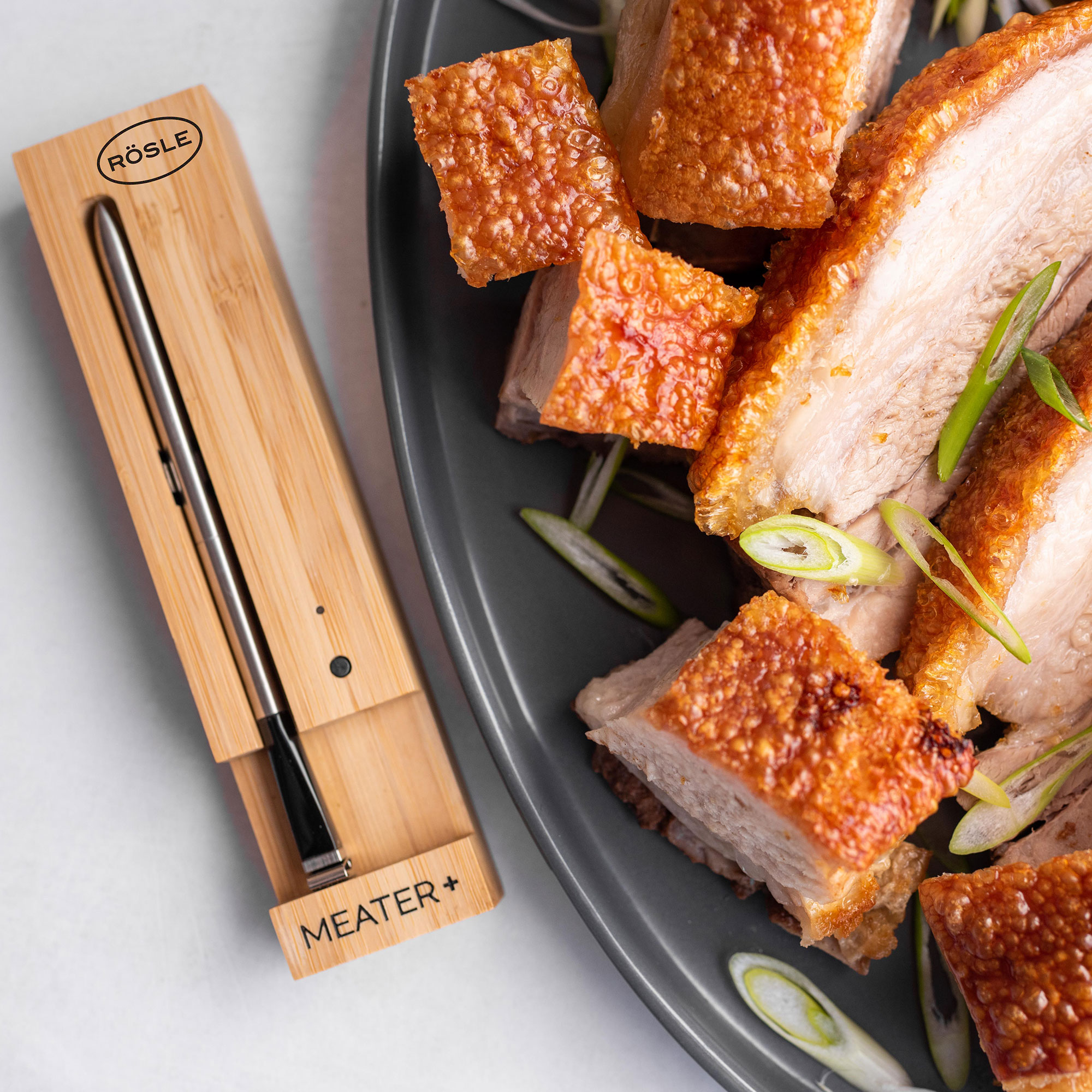 RÖSLE MEATER+ Fleischthermometer und Schweinefleisch MEATER+ Fleischthermometer aus Bambus neben knusprigem Schweinefleisch auf einem grauen Teller,...