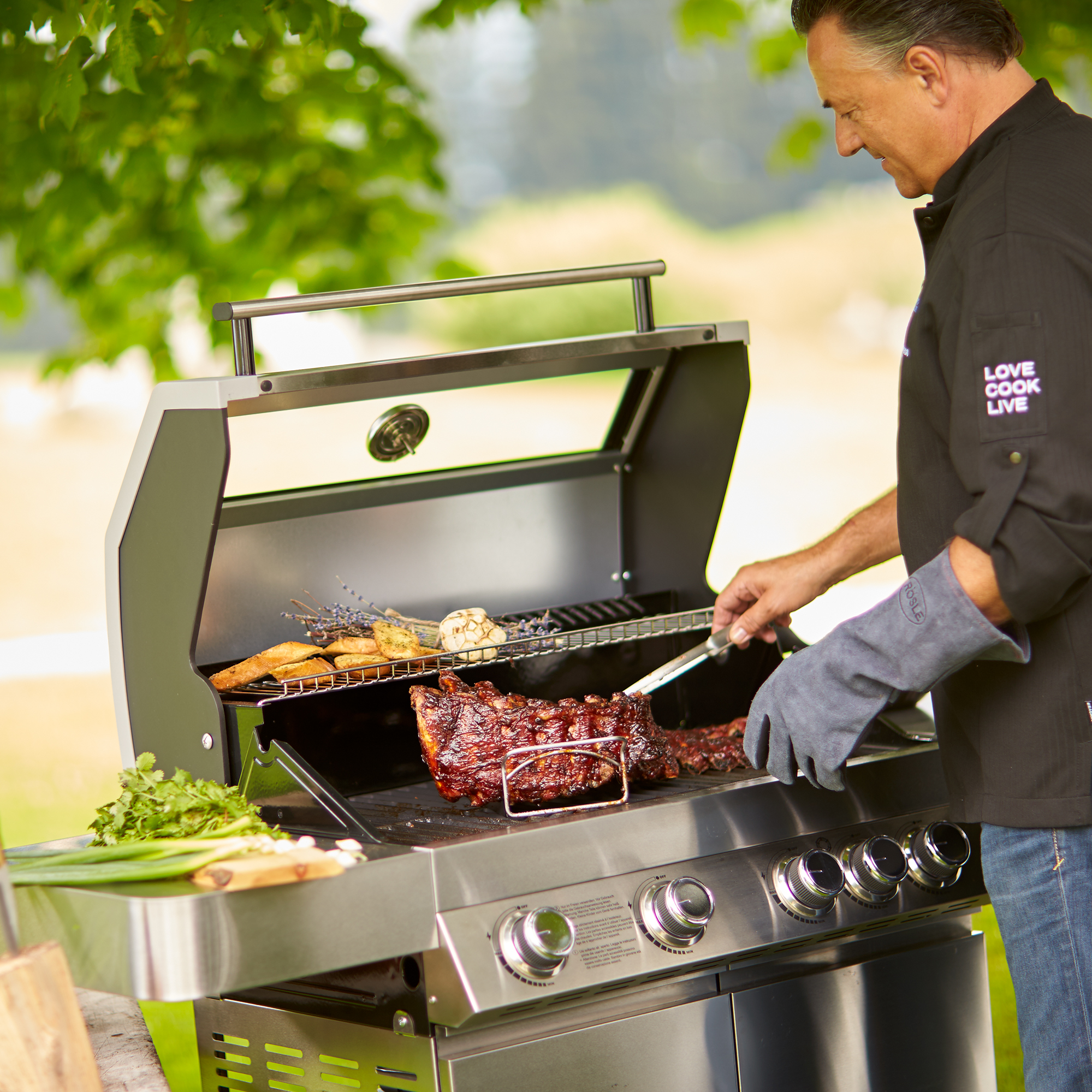 RÖSLE Gasgrill Videro G4 im Einsatz Gasgrill Videro G4 aus Edelstahl mit Grillgut, Person in schwarzer Kochjacke grillt im Freien.