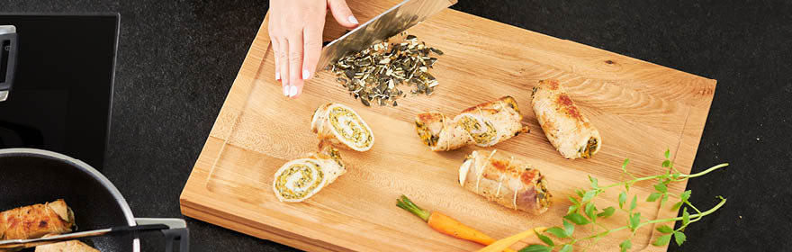 Houten snijplank met gevulde broodjes en pompoenpitten Houten snijplank met een hand die een mes vasthoudt terwijl er gevulde broodjes en pompoenpitten...