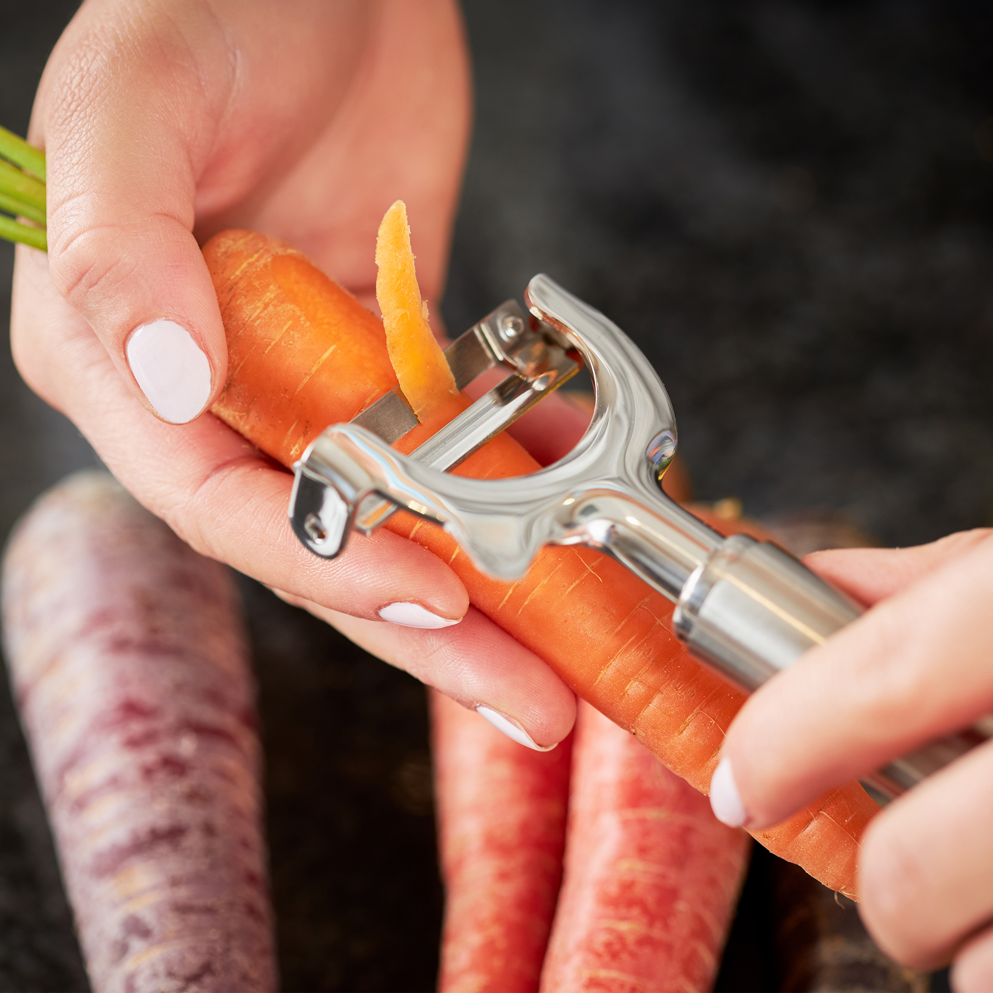 RÖSLE Pendelschäler in Aktion Pendelschäler aus Edelstahl, der eine Karotte schält, mit bunten Karotten im Hintergrund.
