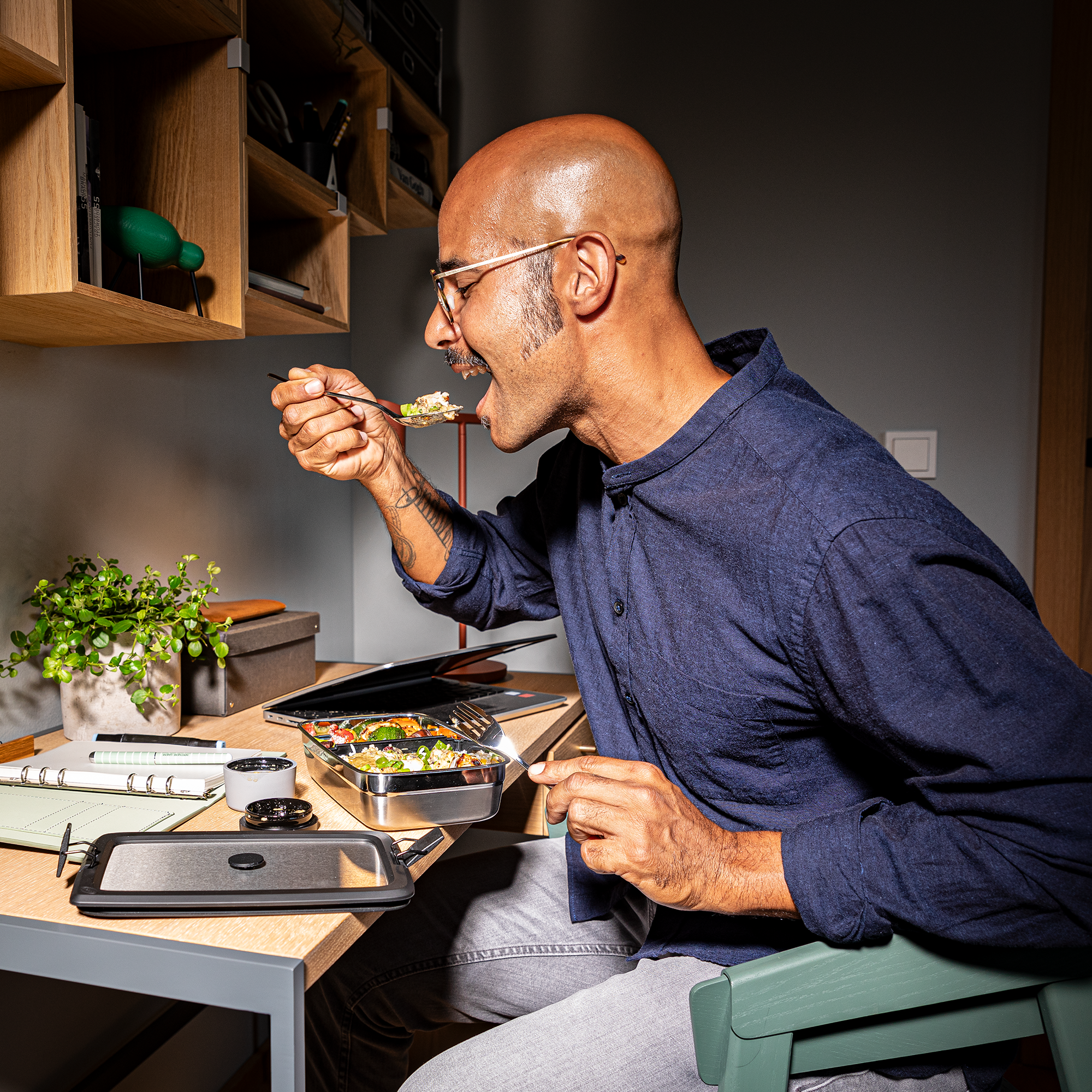 Mann mit Glatze und Brille, der am Schreibtisch aus einer Schüssel mit Essen isst. Im Hintergrund sind Pflanzen und Büromaterial sichtbar.