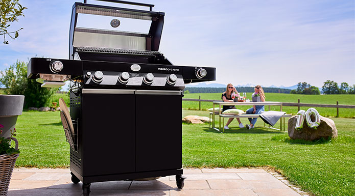 RÖSLE Gasgrill BBQ-Station Videro G4-S NERO Gasgrill mit geöffnetem Deckel im Vordergrund. Dahinter zwei Damen, die mit Weingläser anstoßen.