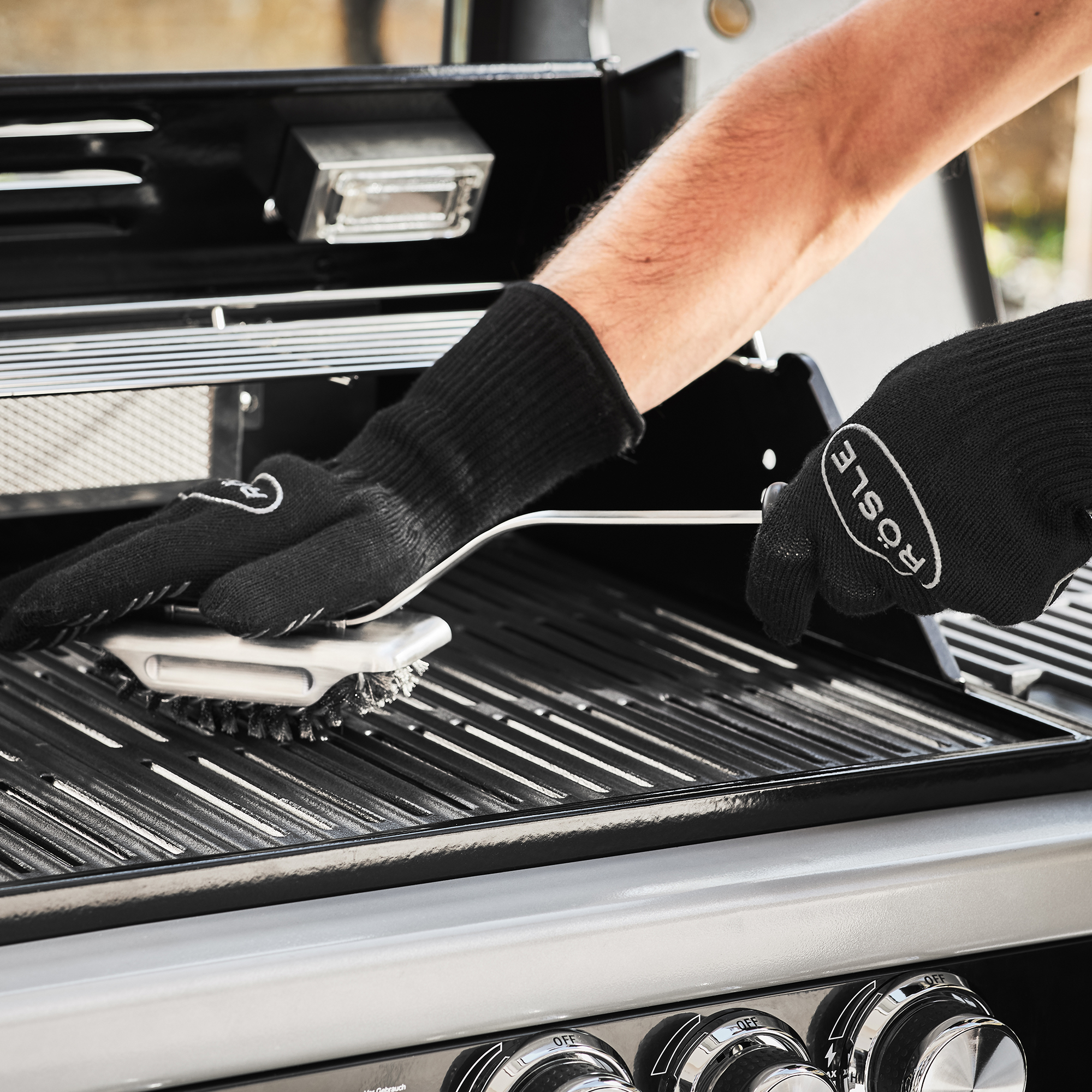 RÖSLE BBQ cleaning brush on grill BBQ cleaning brush is used on a grill grate, hand with black grill glove.