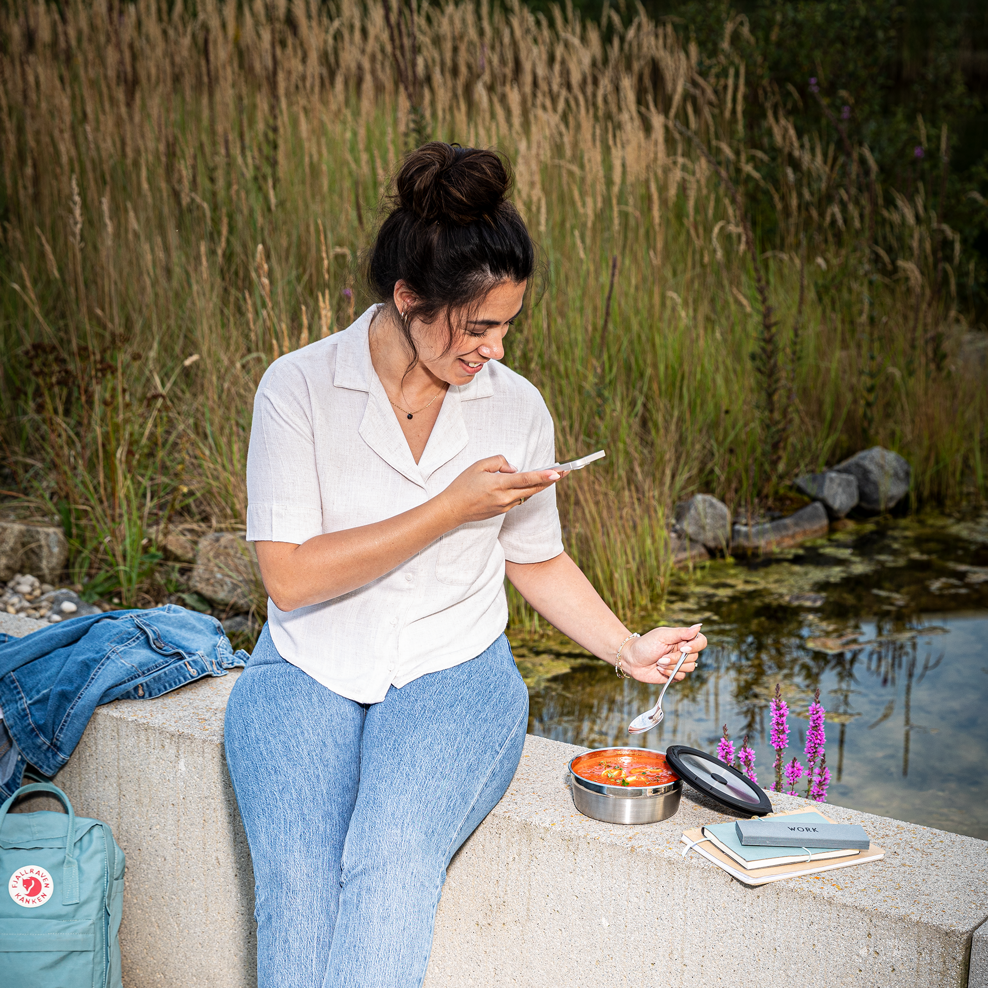 Frau mit dunklem Haar in einem weißen Hemd sitzt am Ufer, isst aus einer Flexx2go Dose und schaut auf ihr Handy.