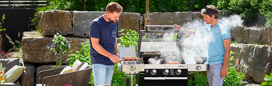 Twee mannen barbecueën buiten met een grill, de rook stijgt op, omringd door planten.