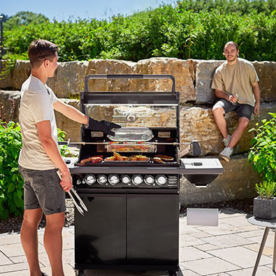 A man stands at the grill with grill tongs in hand and opens the spit basket full of vegetables with the grill glove.