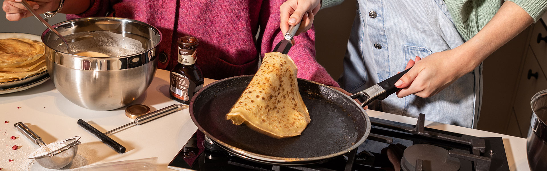 Preparation of crêpes in a RÖSLE pan Two people prepare crêpes in a kitchen, one person turns a crêpe in a pan.