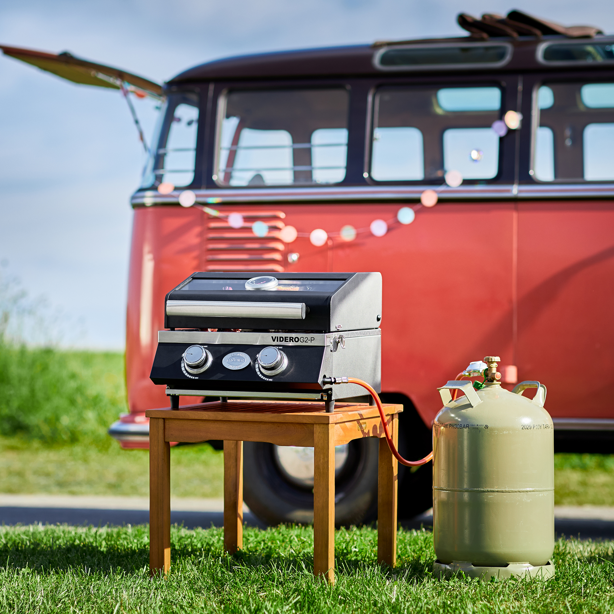 Gasgrill Videro G2-P auf einem Tisch neben einer Gasflasche, im Hintergrund ein Oldtimer-Bus.