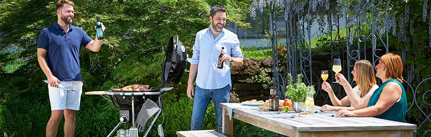 Grillvergnügen im Freien mit RÖSLE Holzkohlegrill Holzkohlegrill im Freien mit zwei Männern, die grillen und Getränke halten, während vier Frauen...