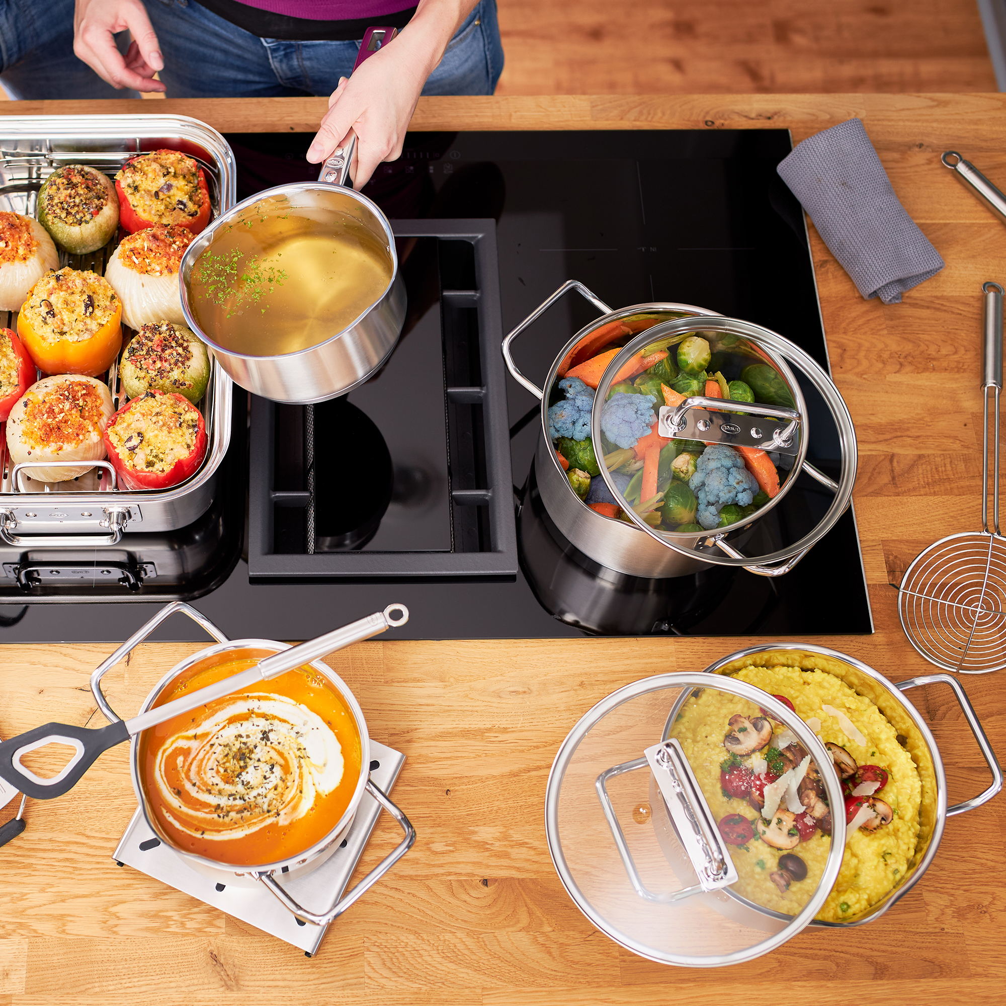 Kochszenario mit RÖSLE Kochtopf und verschiedenen Gerichten Kochtopf mit Gemüse und Brühe auf einem Herd, umgeben von weiteren Kochgeräten und Speisen.