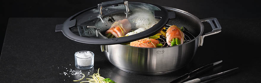 Roestvrijstalen braadslee met glazen deksel, gevuld met groenten en zalm, op een zwarte achtergrond.