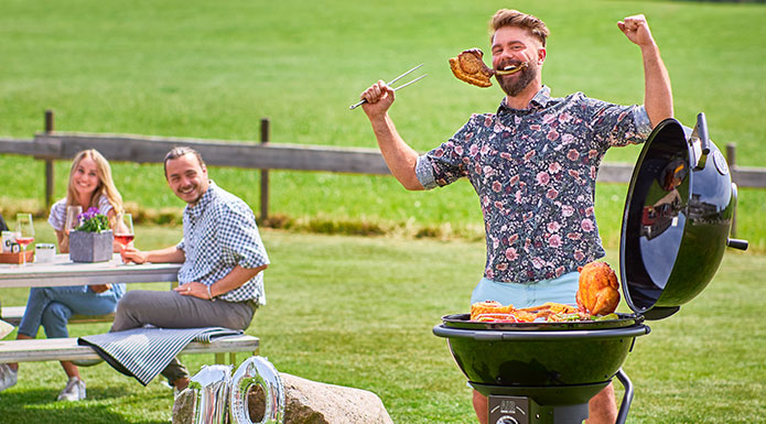 Grillmeister Mann am Kugelgrill jubelt mit einem Stück Fleisch im Mund.