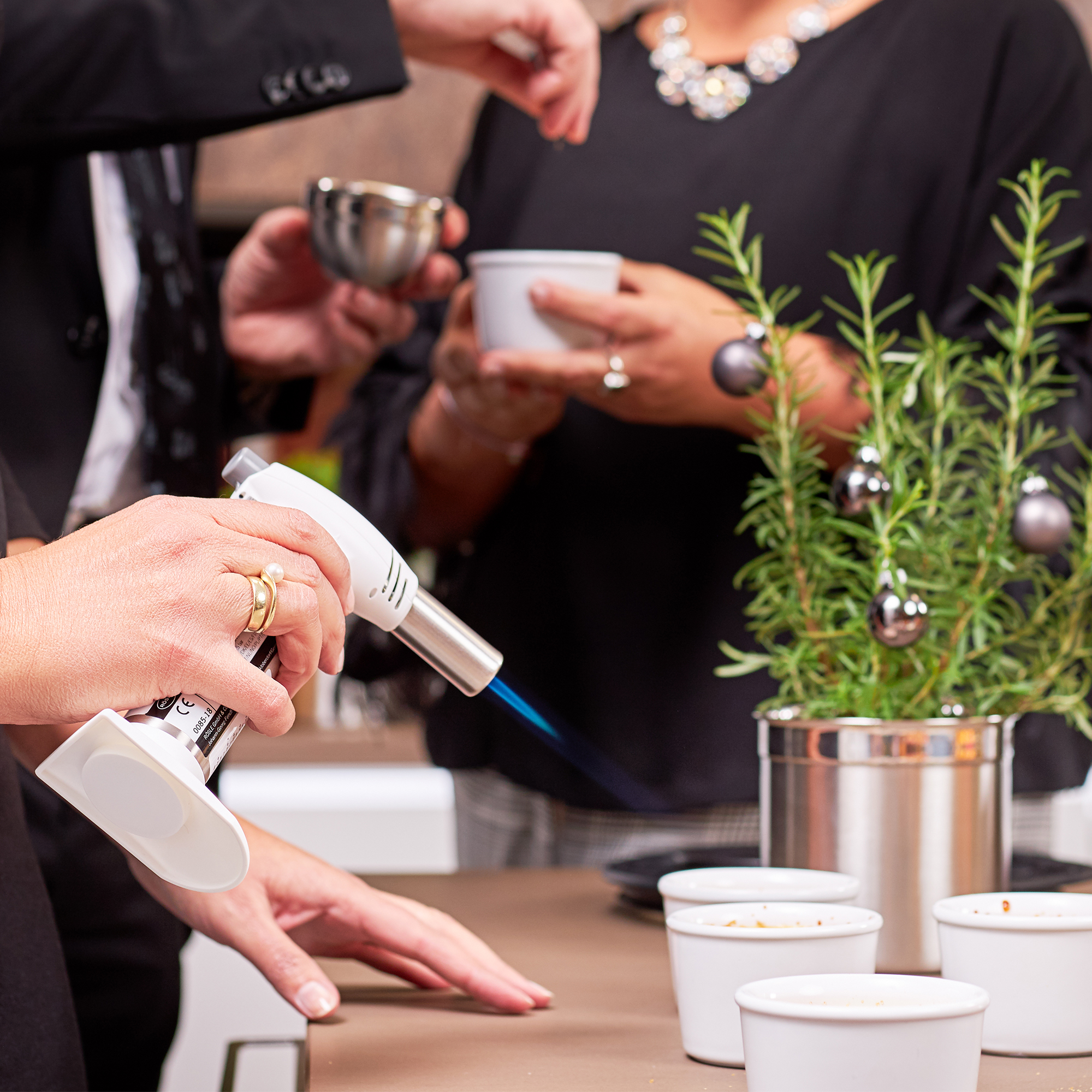 RÖSLE Flambierbrenner in Nutzung Flambierbrenner in Aktion, gehalten von einer Person, während andere Personen in der Hintergrund...