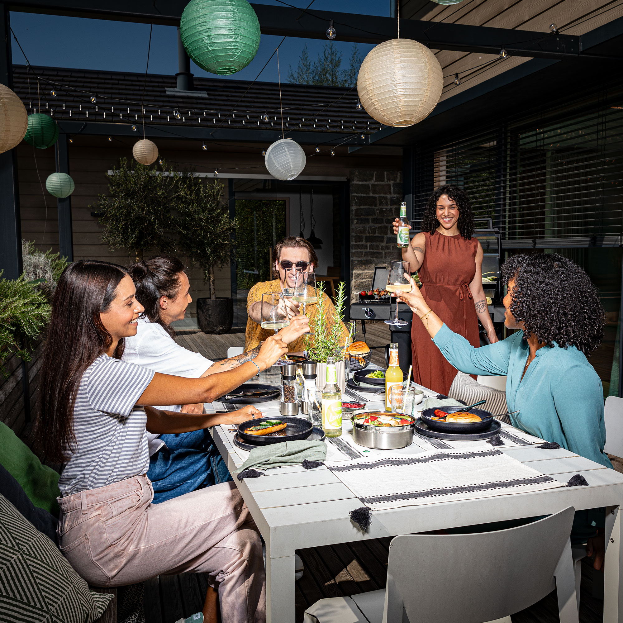 Gutschein BBQ Freunde sitzen zusammen am Tisch auf der Terrasse und stoßen gemeinsam an