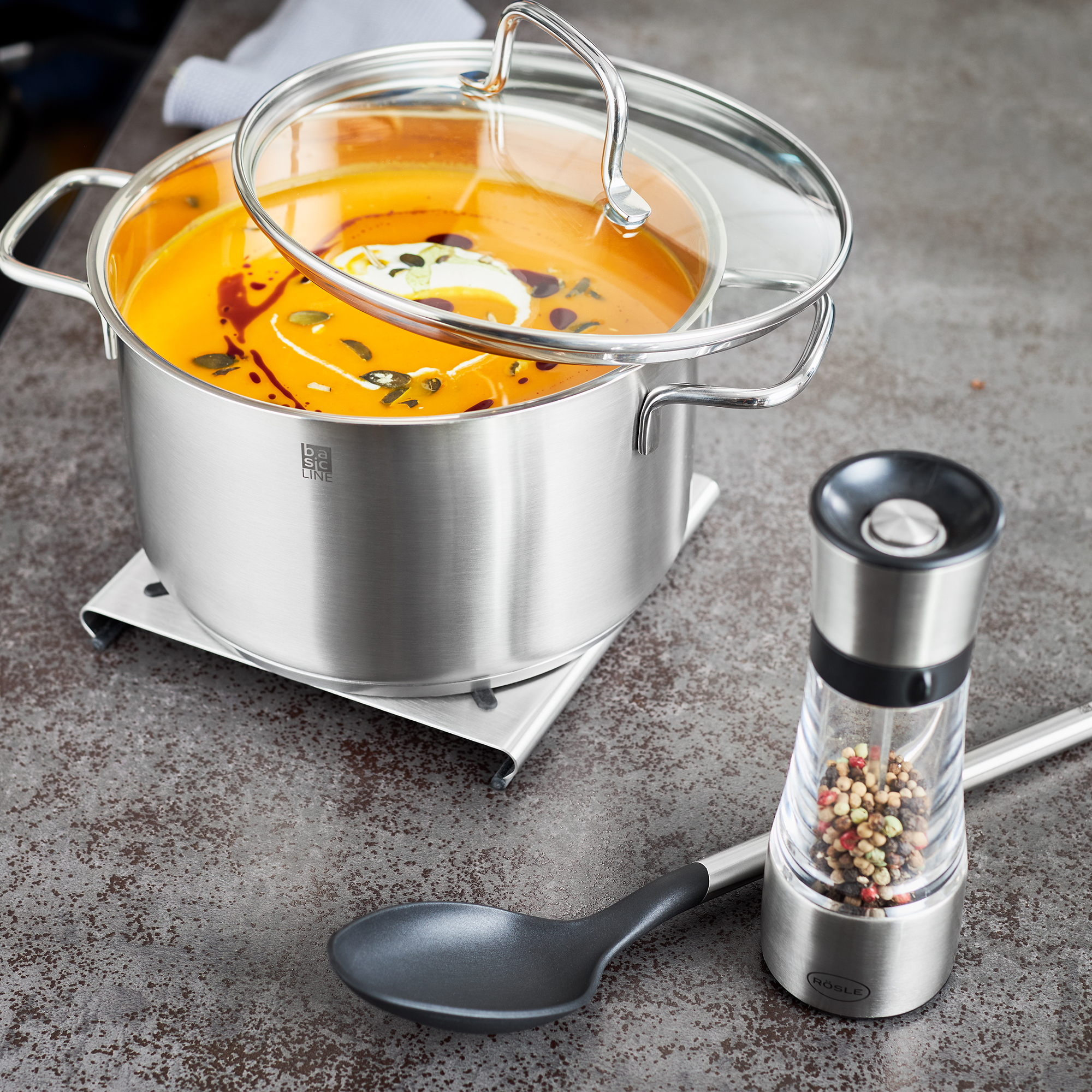 Edelstahl Topfuntersetzer mit Kochtopf und Zubehör Topfuntersetzer aus Edelstahl unter einem Kochtopf mit Suppe und Glasdeckel, daneben eine...