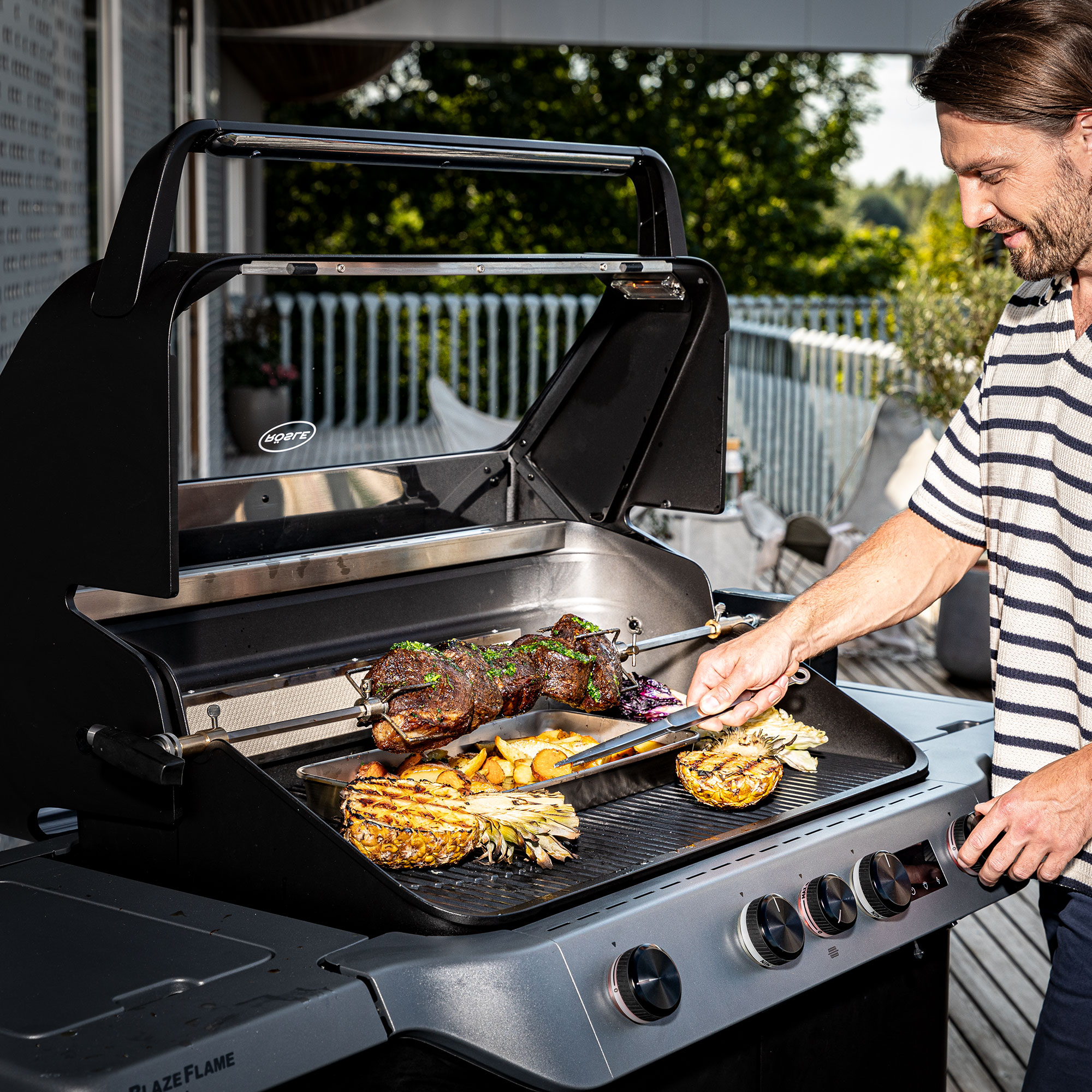 Drehspieß mit Grillgut und Gemüse auf einem Grill, Mann in gestreiftem Shirt bedient den Grill.