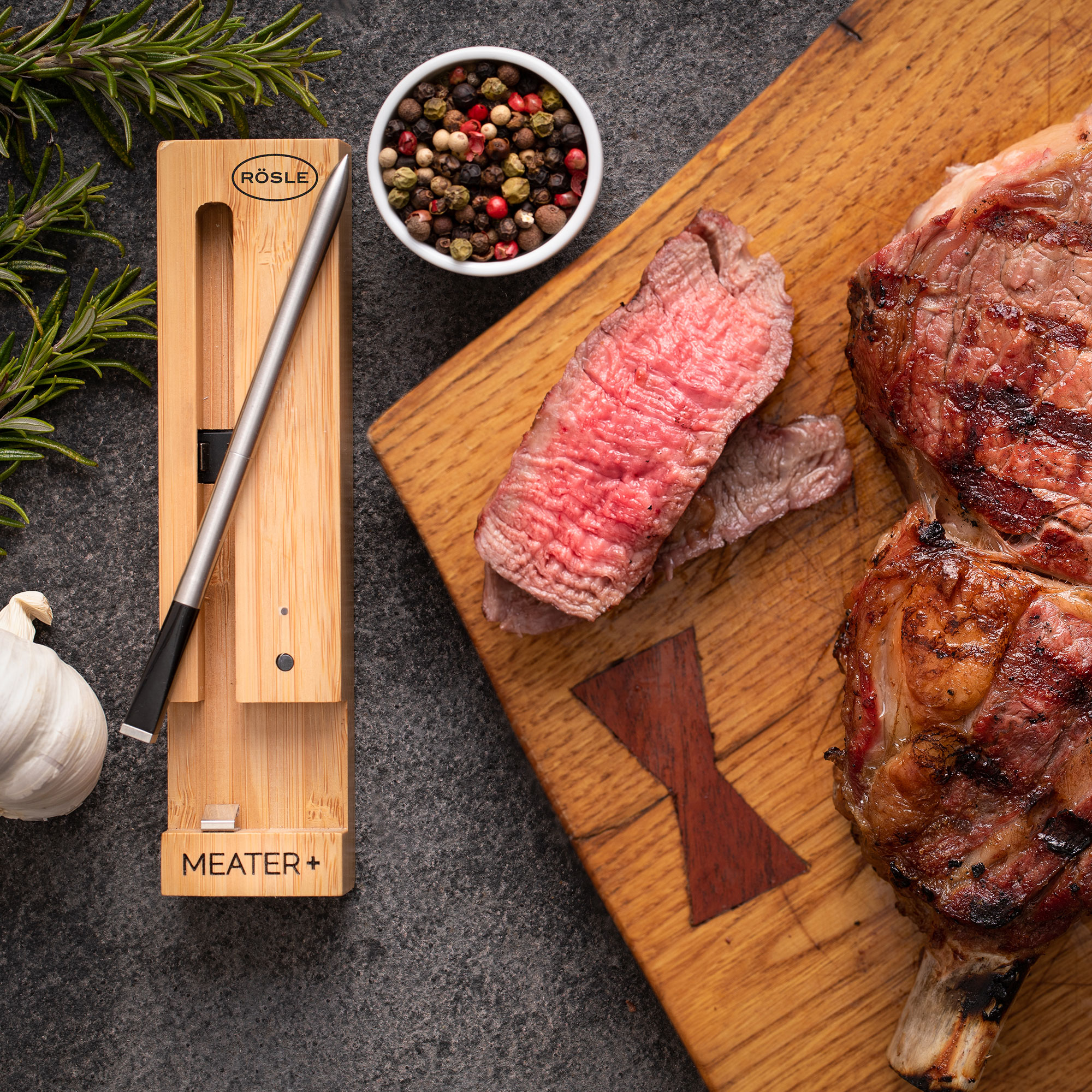 RÖSLE Meater Plus Holzthermometer mit gegrilltem Fleisch Meater Plus, ein Holzthermometer für Fleisch, neben gegrilltem Fleisch und Gewürzen auf einem...