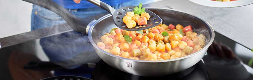 BASIC LINE Stainless steel frying pan with colourful vegetables, plastic cooking spoon, steam rises