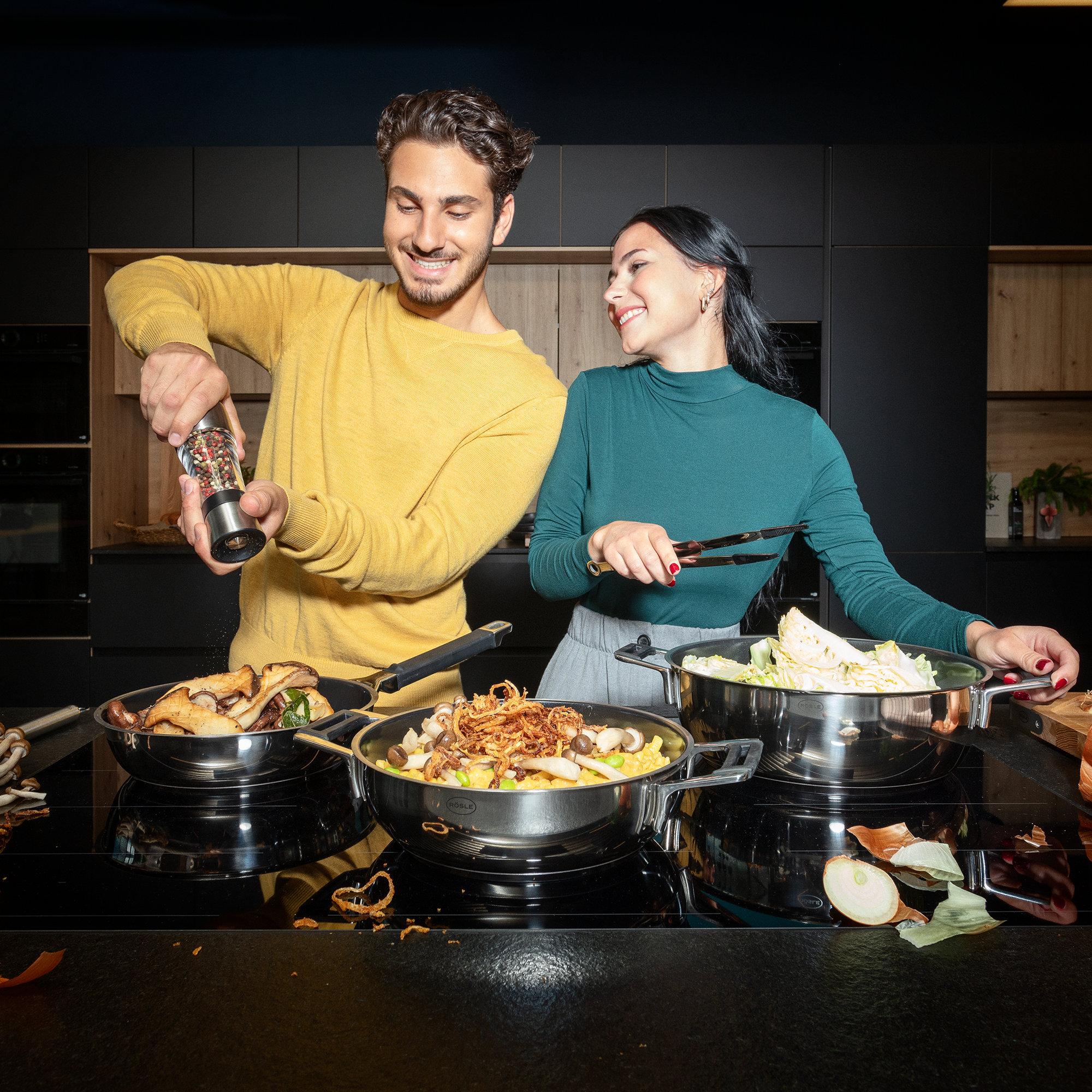 Kochszene mit RÖSLE Topf-Set Zwei Personen in einer modernen Küche, die gemeinsam mit Kochgeschirr kochen. Der Mann würzt...