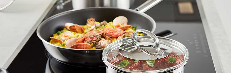 RÖSLE Pfanne mit Meeresfrüchten und Gemüse Pfanne mit Meeresfrüchten und Gemüse auf dem Herd, daneben ein Topf mit Deckel und Kräutern.