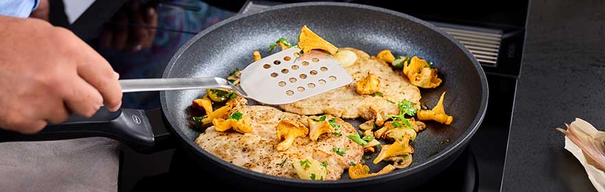 CADINI Pfanne mit gebratenen Pilzen und Fleischstücken, eine Hand hält einen Pfannenwender.