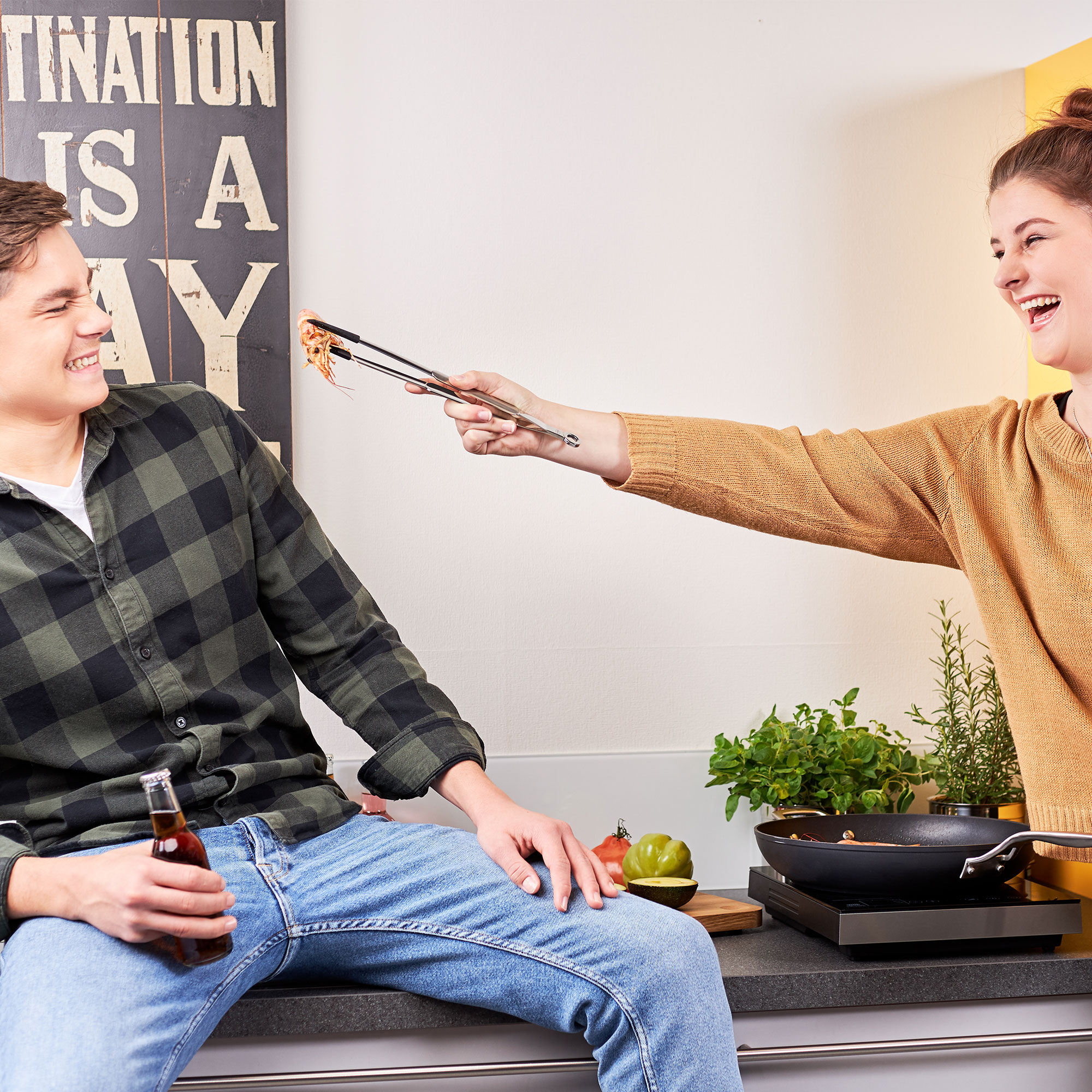 Two people in the kitchen with RÖSLE needle nose tongs Two people in the kitchen, one person hands food to the other, who is sitting relaxed, with...