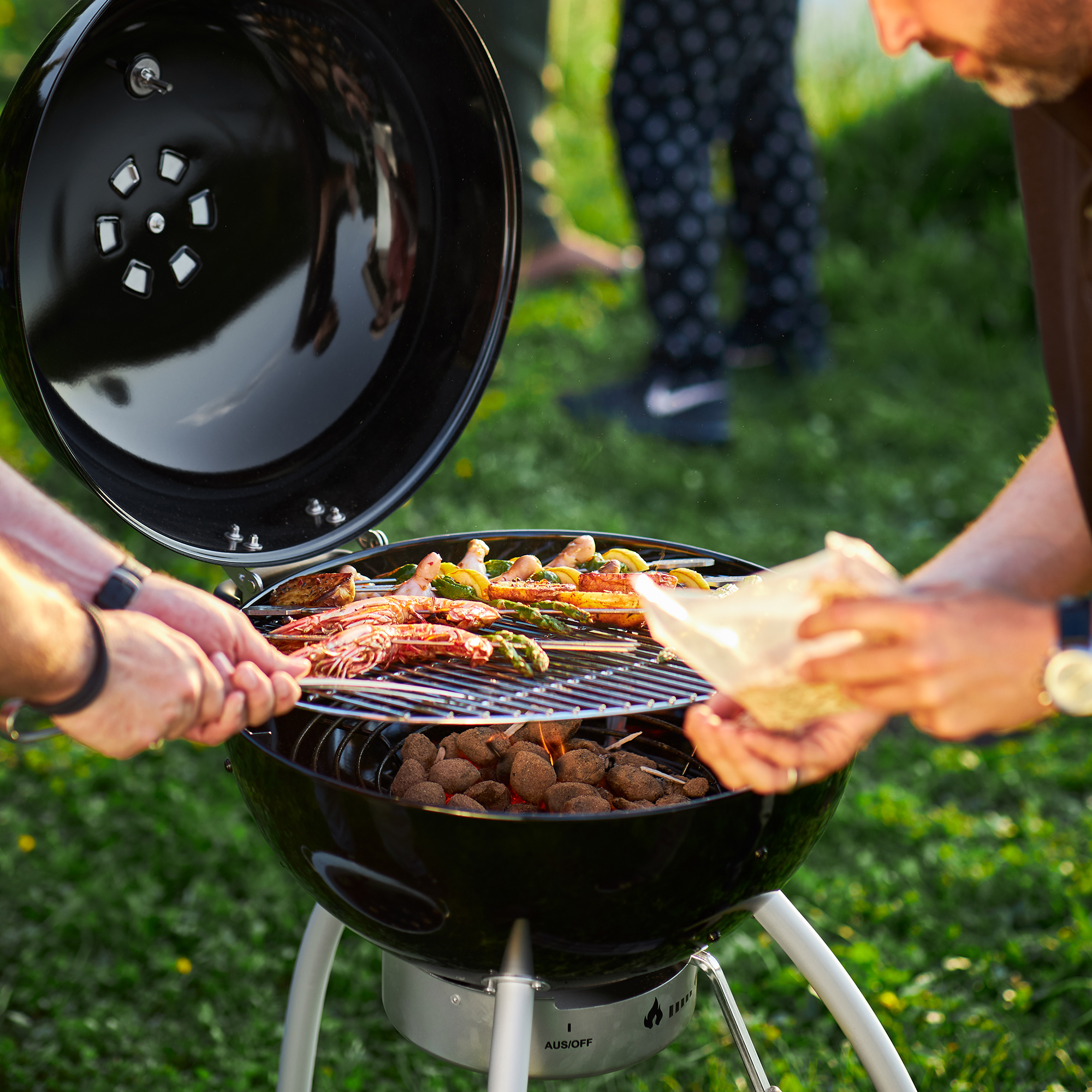 Grill mit RÖSLE Kohlezange und Grillgut Kohlezange in Verwendung, Grill mit Meeresfrüchten und Gemüse, grüne Wiese im Hintergrund