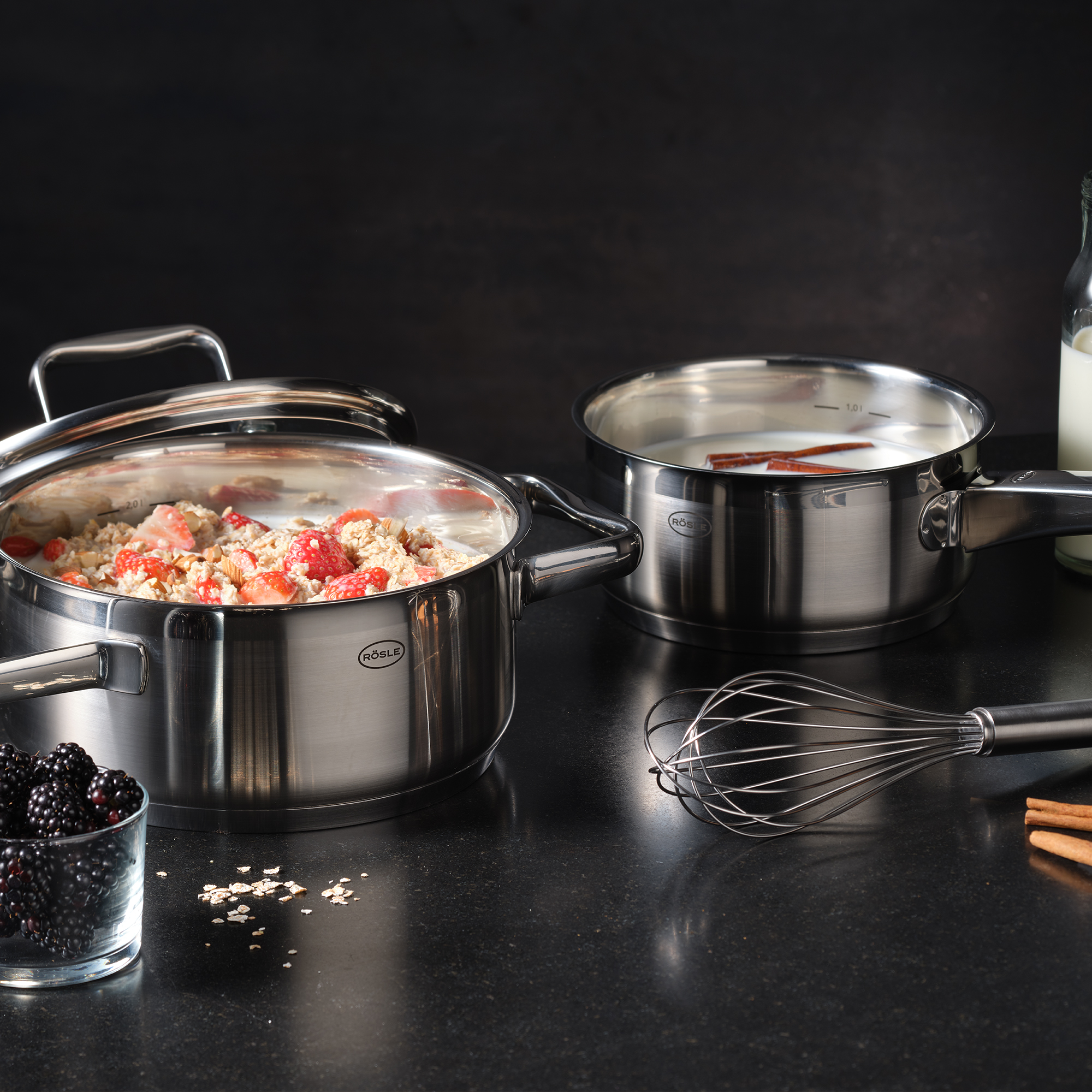 RÖSLE saucepans and whisks Stainless steel cooking pots with lids, filled with a mixture of oatmeal and fruit, next to...