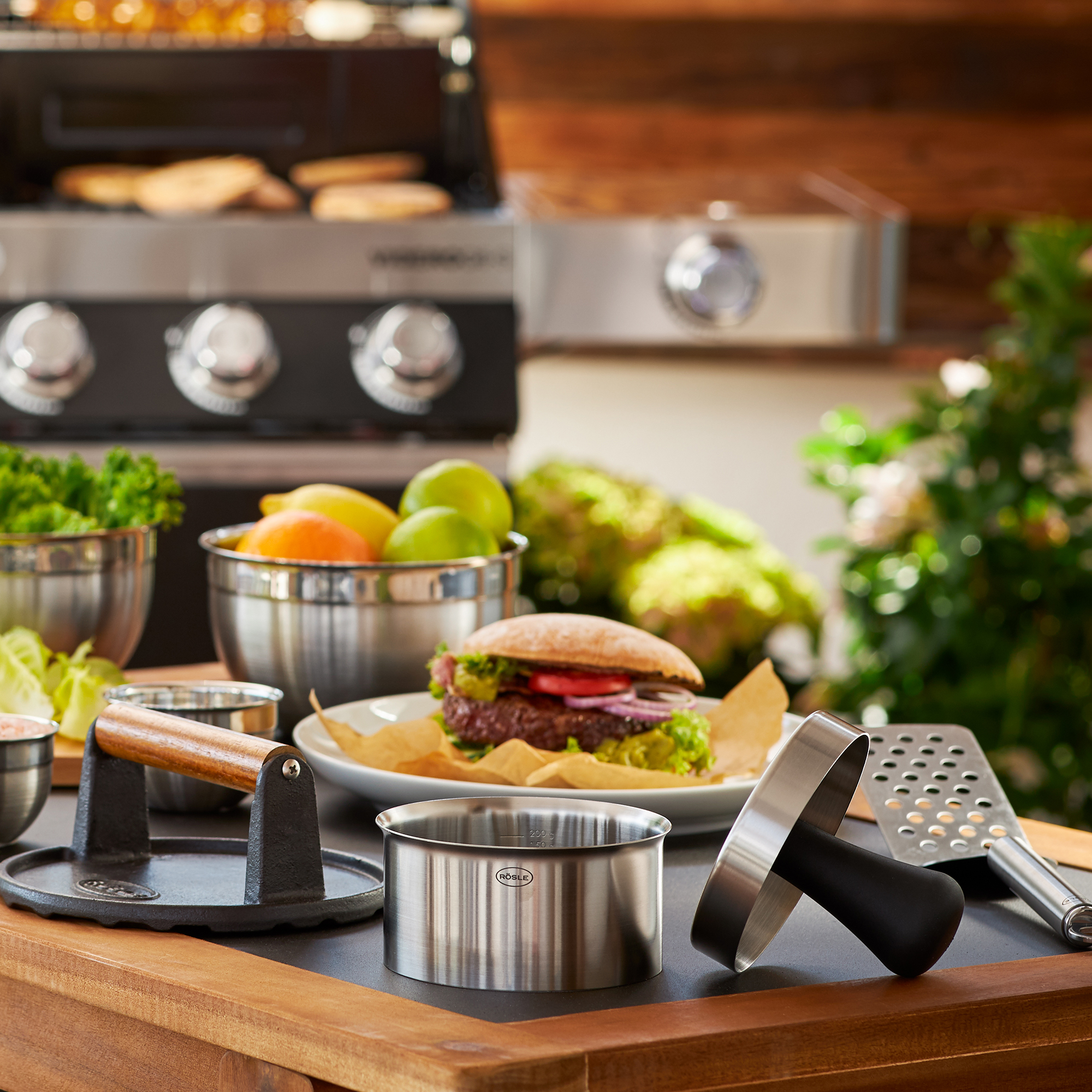 RÖSLE burger press and barbecue accessories Stainless steel burger press with wooden handle, surrounded by fresh ingredients and barbecue...