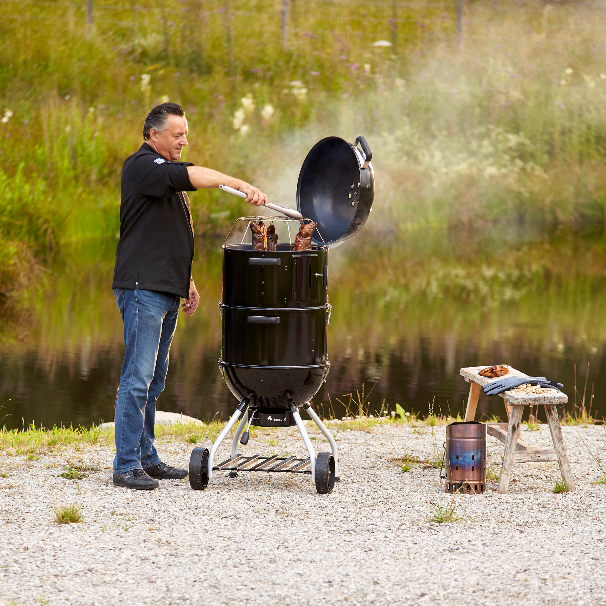RÖSLE Holzkohle-Wasser-Smoker im Einsatz Holzkohle-Wasser-Smoker in Schwarz, ein Mann öffnet den Deckel, Rauch steigt auf, im Freien.