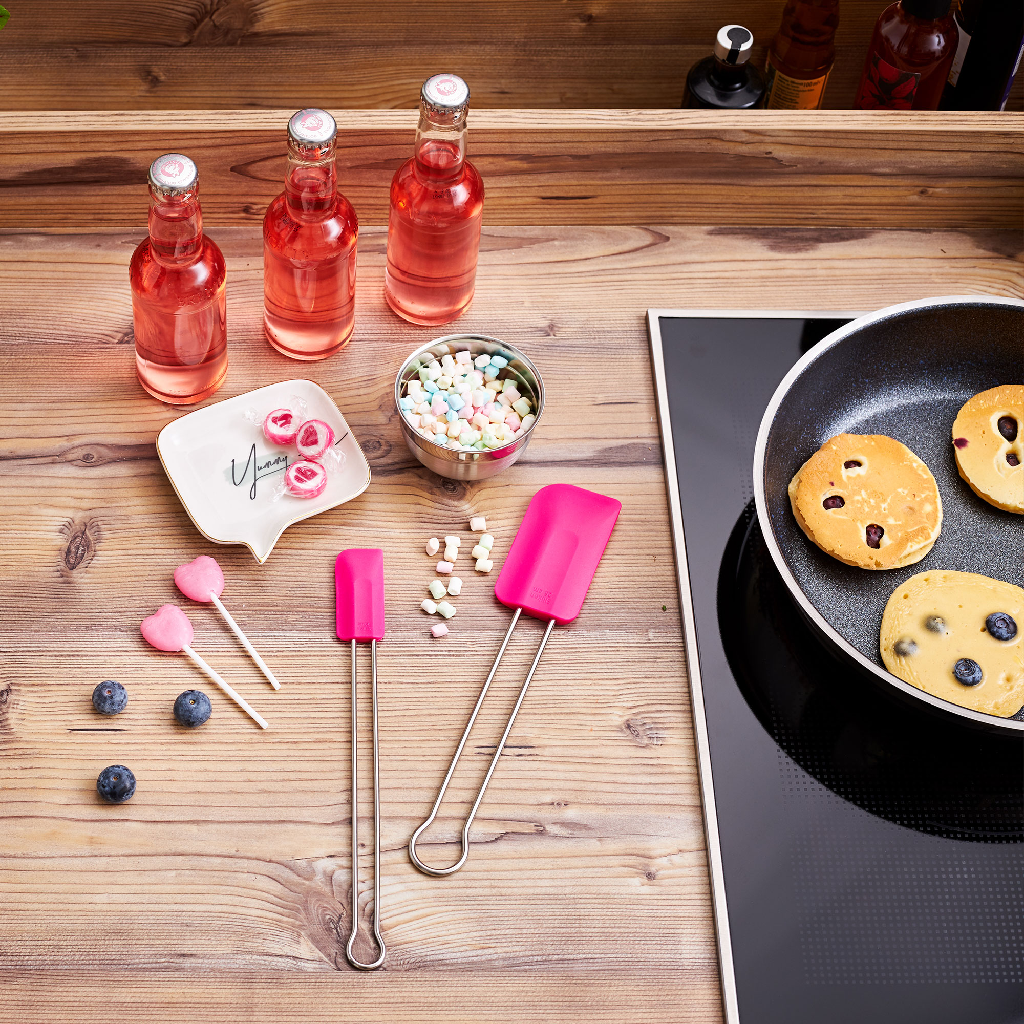 RÖSLE Teigschaber 26cm in Pink Teigschaber in Pink, neben bunten Süßigkeiten, Getränken und Pfannkuchen auf einer Arbeitsfläche.