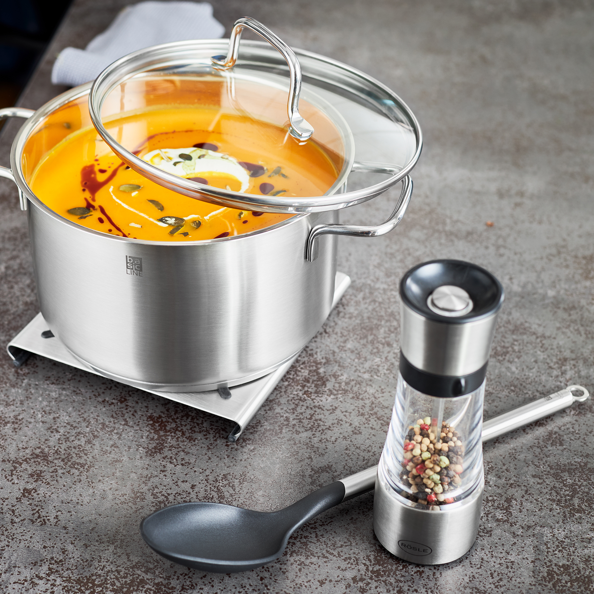 RÖSLE Gewürzmühle und Suppentopf Gewürzmühle aus Edelstahl neben einem Topf mit orangefarbener Suppe und einem Kochlöffel.