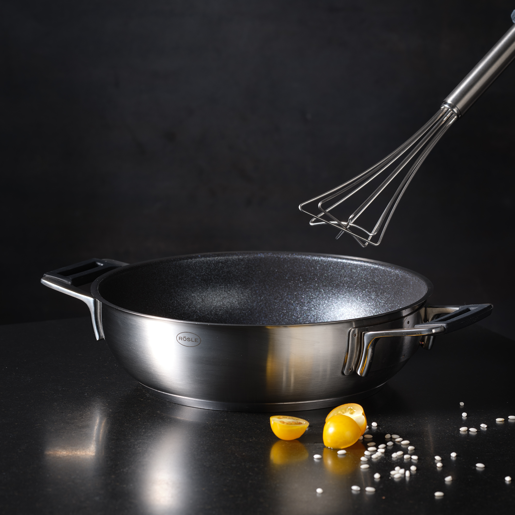 RÖSLE Gourmetbesen und Pfanne Gourmetbesen aus Edelstahl, 27 cm, neben einer Pfanne mit Antihaftbeschichtung und Zitronenstücken.