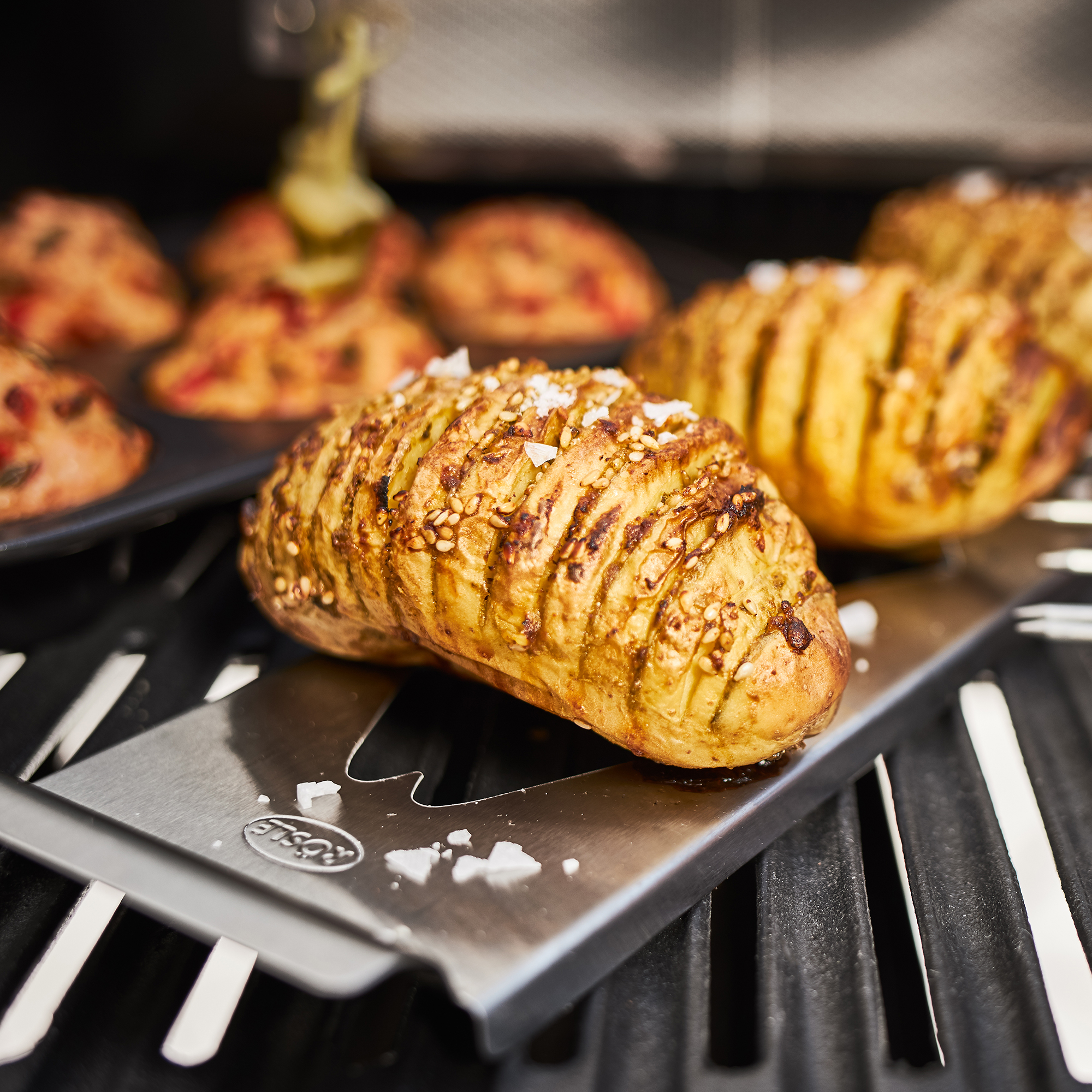RÖSLE Kartoffelhalter mit gegrillter Kartoffel Kartoffelhalter mit einer gegrillten Kartoffel, die Rillen aufweist, auf einem Grillrost.