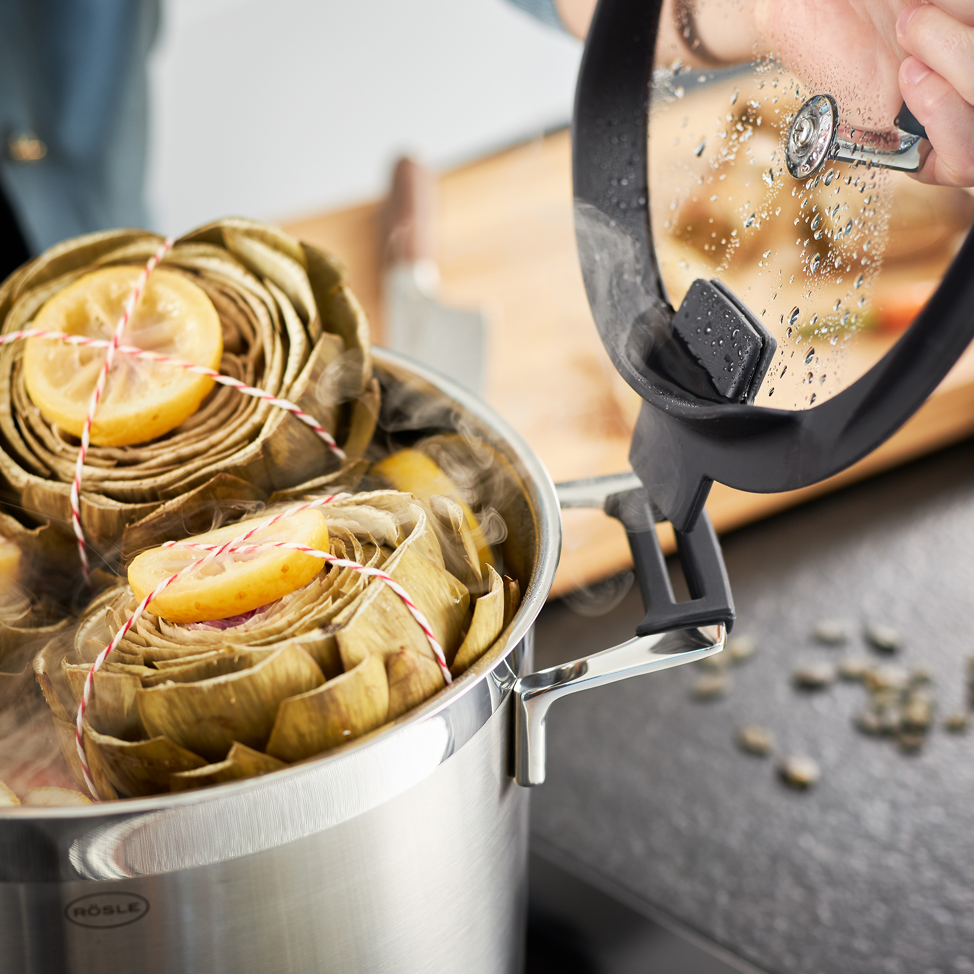 RÖSLE Kochtopf 24cm hoch mit Artischocken Kochtopf aus Edelstahl mit Deckel, gefüllt mit Artischocken und Zitronenscheiben, Dampf entweicht.