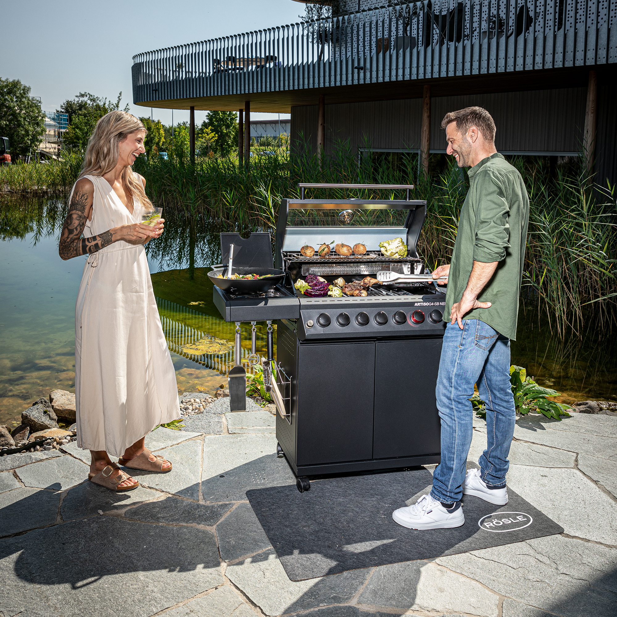 RÖSLE Artiso G4-SB Grill im Freien Artiso G4-SB Grill in Schwarz, im Freien, mit einem Mann und einer Frau, die grillen und Getränke...