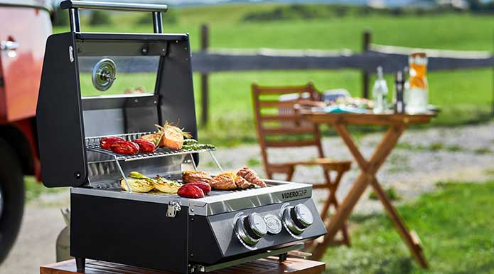 Videro G2-P with open lid and various grilled food in the foreground, small table and chair behind.