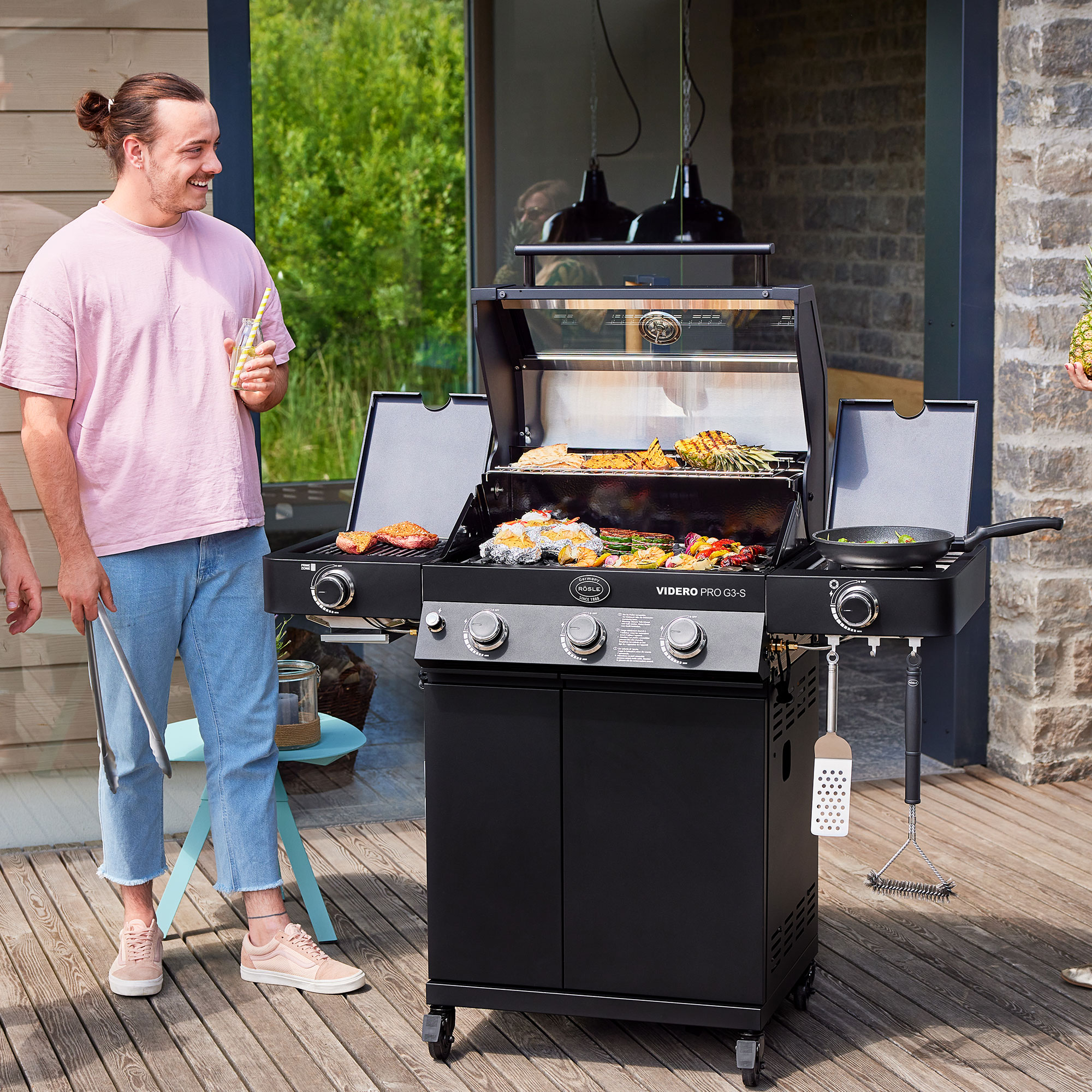 RÖSLE Gasgrill Videro G3-S PRO Gasgrill Videro G3-S PRO in schwarz, mit Grillgut auf dem Rost, zwei Personen stehen daneben.