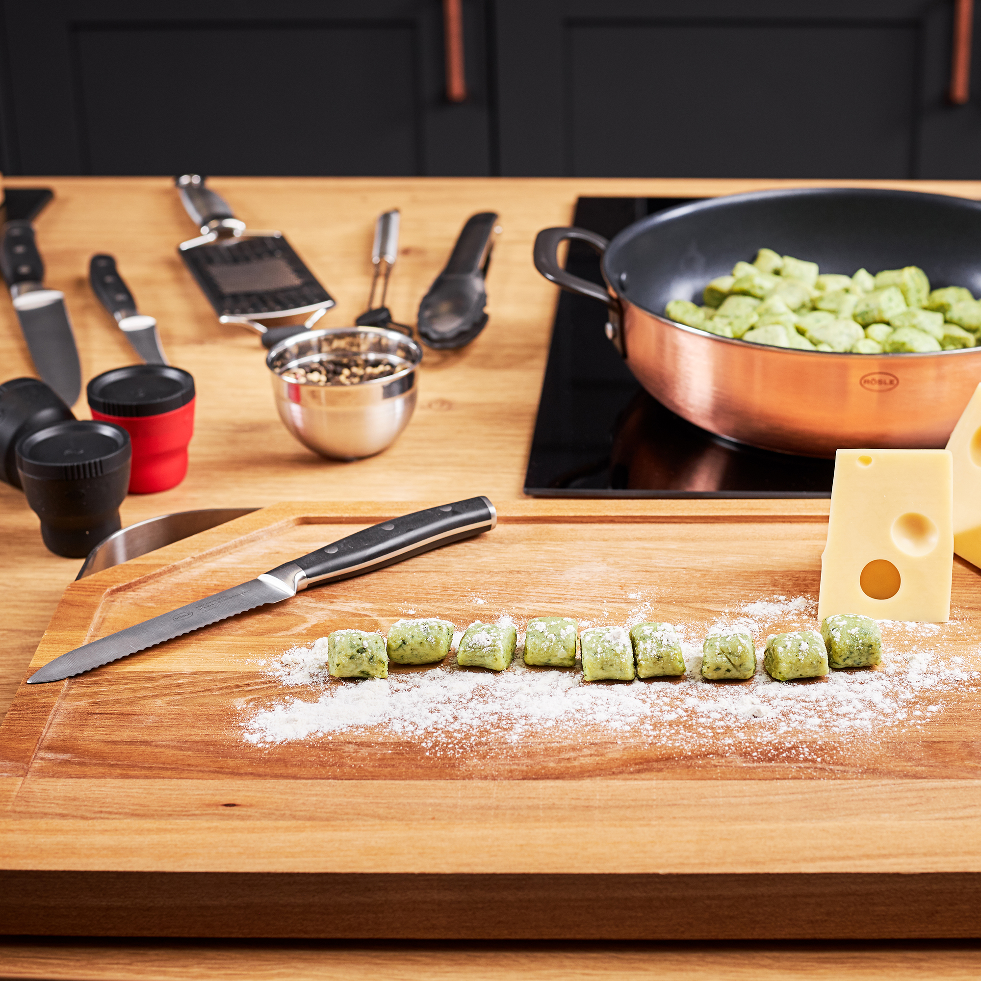 RÖSLE Universalmesser auf Holzbrett mit Gnocchi Universalmesser, 13 cm, auf einem Holzbrett mit Gnocchi und Käse, im Hintergrund Küchengeräte.