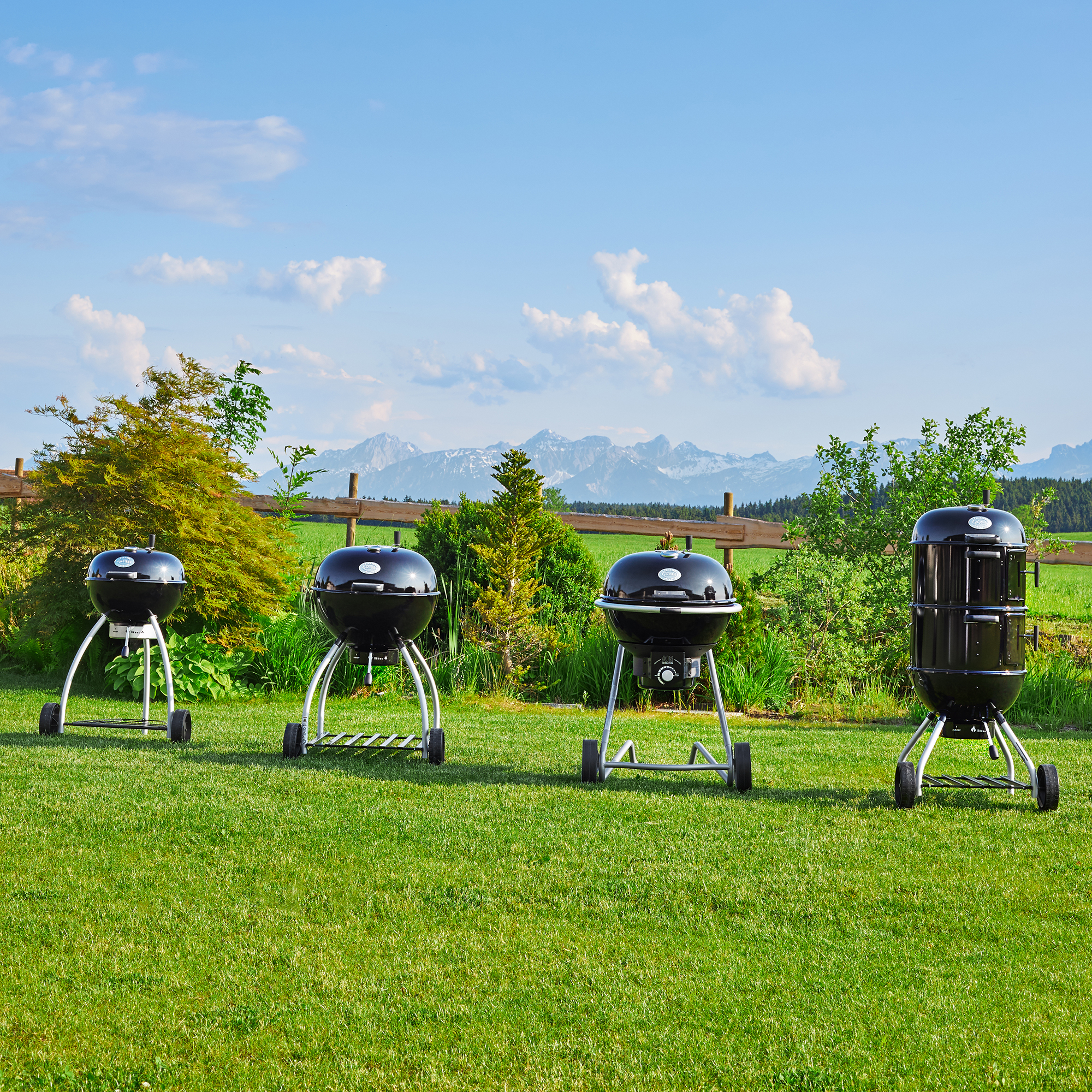 RÖSLE Holzkohlegrills und Wasser-Smoker Holzkohlegrills und Wasser-Smoker in schwarz, auf einer Wiese mit Bergen im Hintergrund.