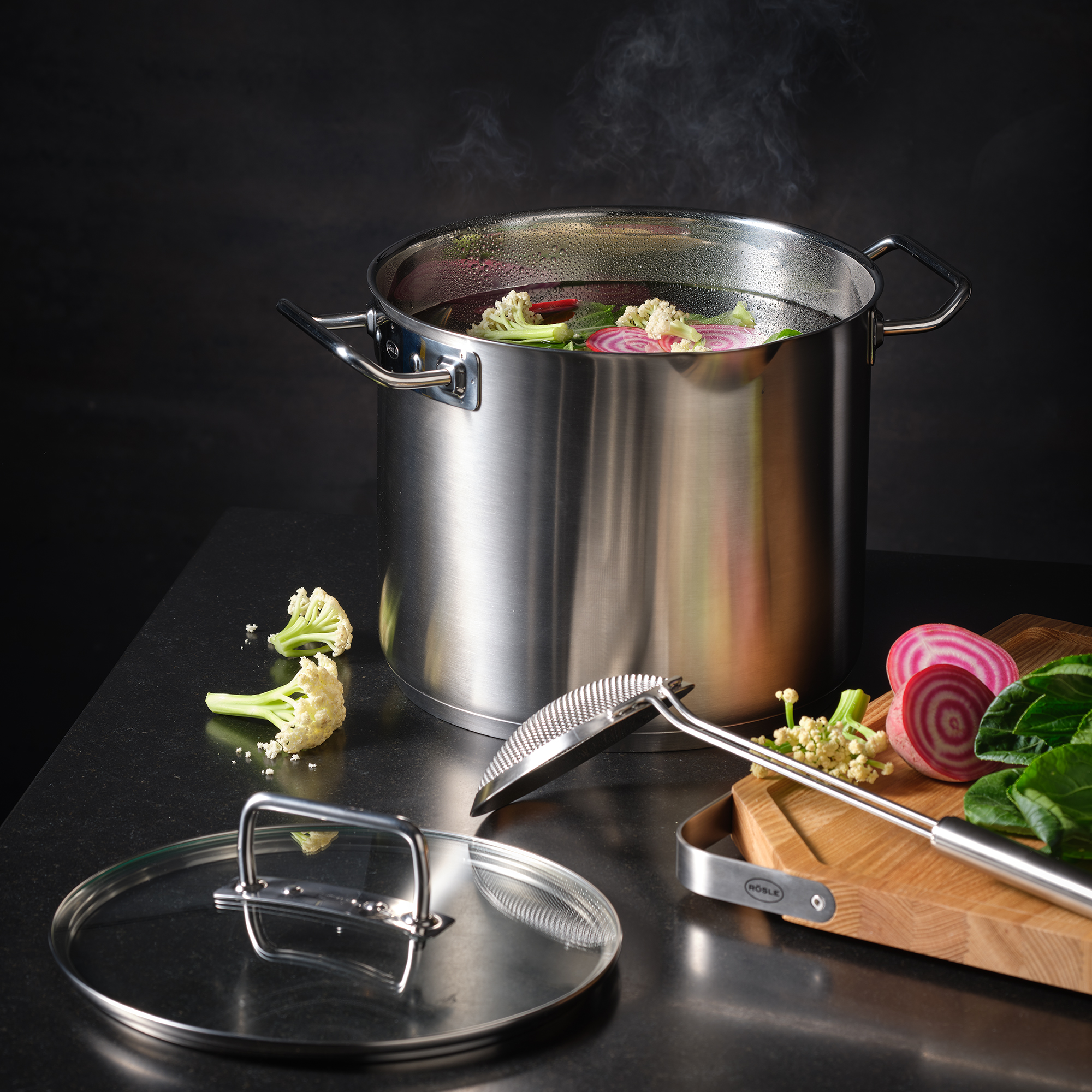 RÖSLE Edelstahl Gemüsetopf 24cm Gemüsetopf aus Edelstahl mit Gemüse und Deckel, neben einem Holzbrett und einem Küchengerät.