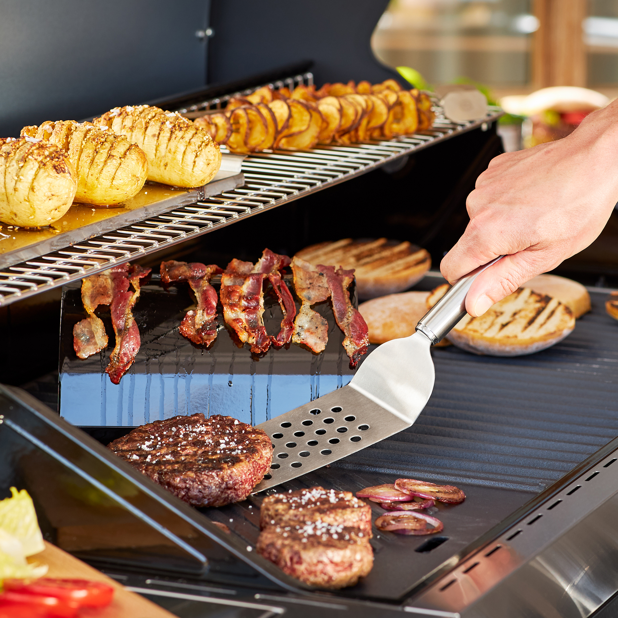 RÖSLE burger turner on the grill Stainless steel burger turner turning burgers on a grill, with grilled vegetables and bacon...