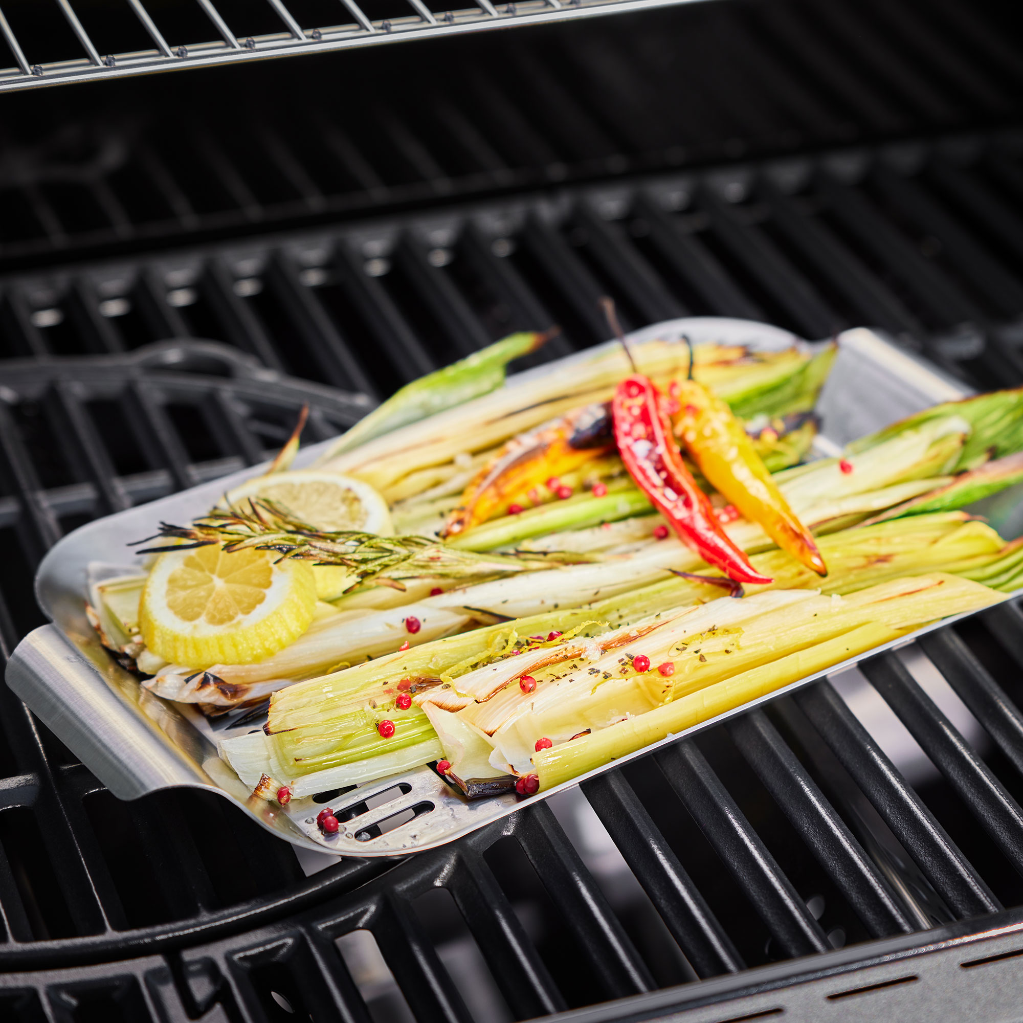 Gemüse-Schale aus Edelstahl mit Löchern, gefüllt mit gegrilltem Gemüse und Zitronenscheiben auf einem Grillrost.