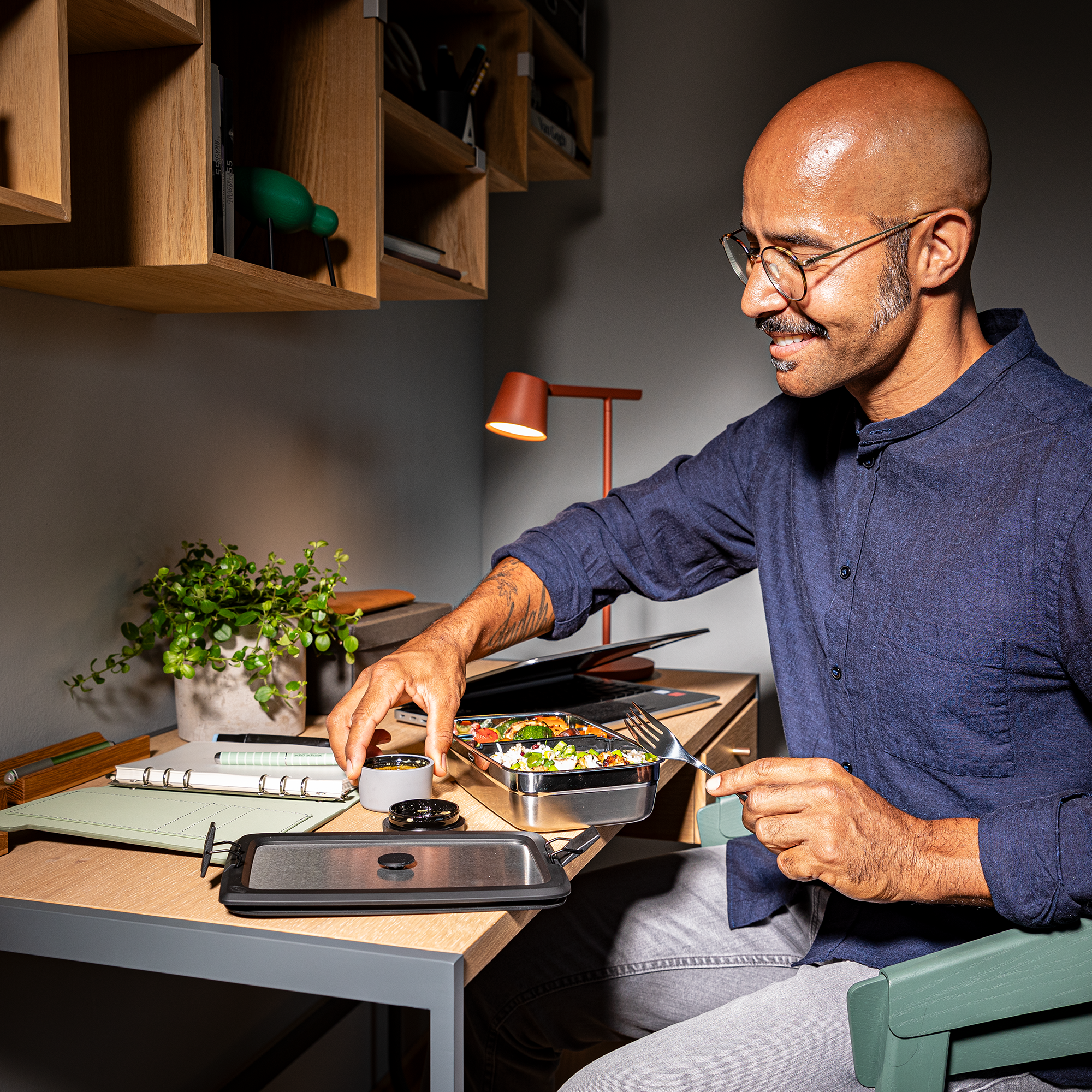 Mann mit Brille, der eine Edelstahl-Lunchbox mit einem Deckel öffnet, während er an einem Schreibtisch sitzt.